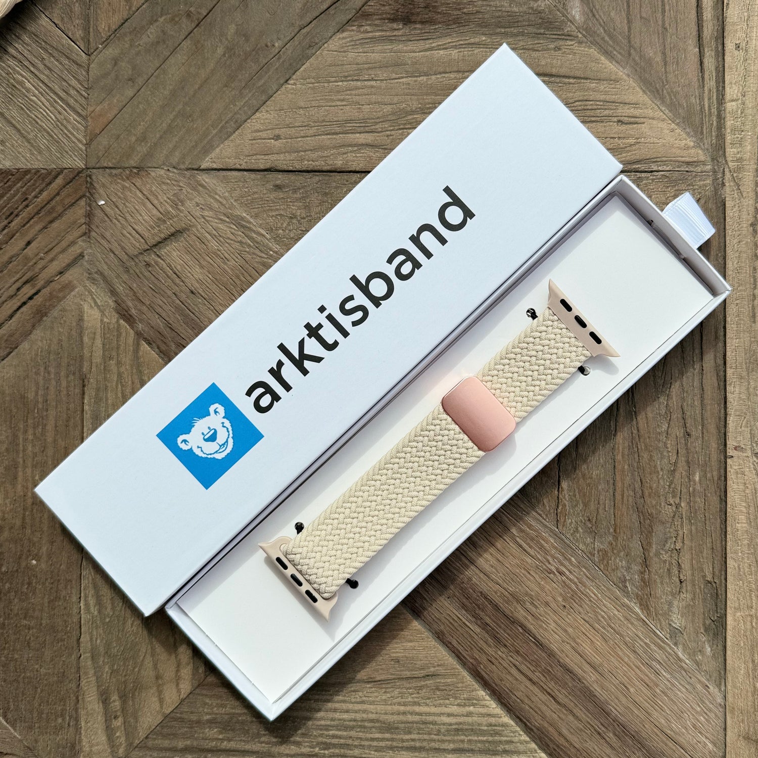 Das arktisband Geflochtene Apple Watch Armband „Casual“ mit Magnetverschluss von arktisband mit rosa Verbindern und Schlaufe wird in einer offenen weißen „arktisband“-Box mit einem blauen Bärenlogo auf einer Holzoberfläche gezeigt.