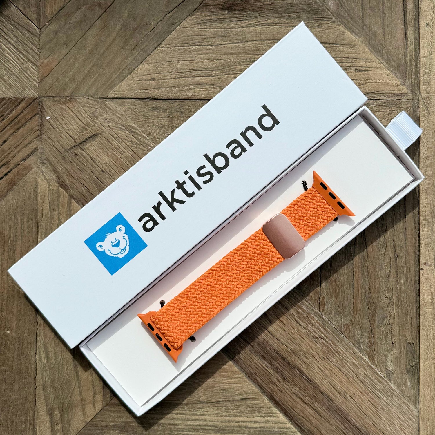 Das arktisband Geflochtene Apple Watch Armband „Casual“ mit Magnetverschluss von arktisband in Orange liegt in einer offenen weißen arktisband Box auf einer Holzoberfläche.