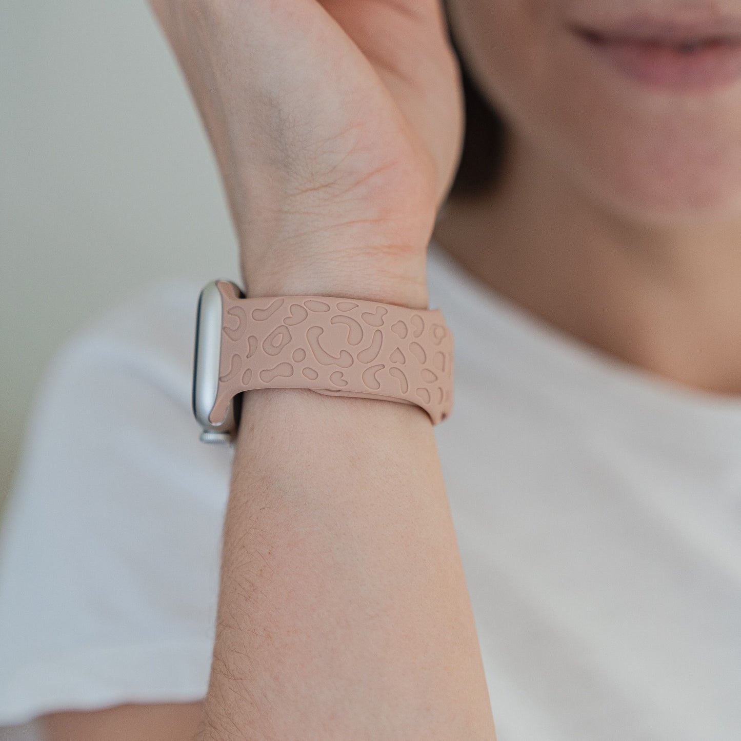 Eine Person trägt ein Apple Watch-kompatibles Arktisband Apple Watch Armband „Leopard“ in Hellrosa mit erhabenem Muster. Sie trägt ein schlichtes weißes Hemd und lächelt leicht, wobei ihr Gesicht teilweise sichtbar ist.