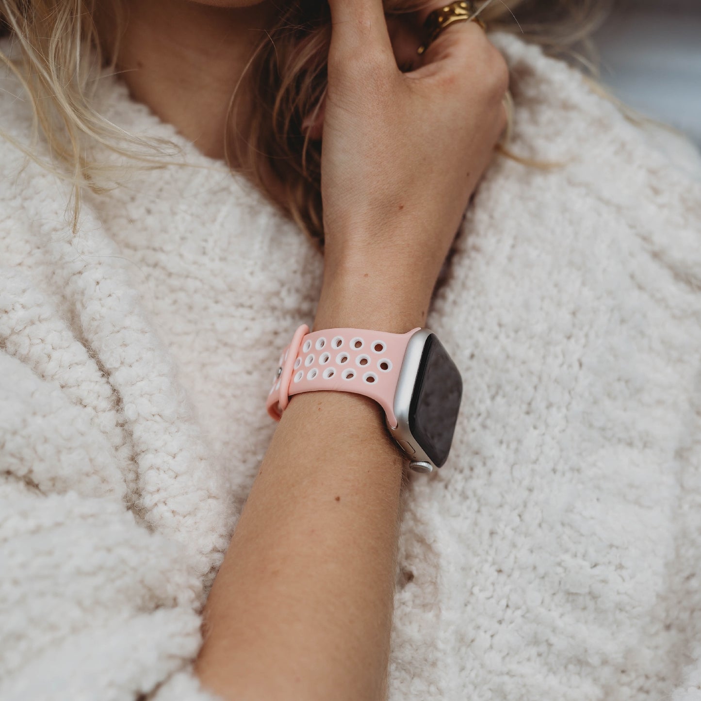Eine Person in einem weißen Strukturpullover trägt ein arktisband Apple Watch Silikonarmband „Energy“ von arktisband in Pink und Weiß am Handgelenk. Ihre Hand liegt nah am Gesicht, teilweise sind blonde Haare zu sehen.