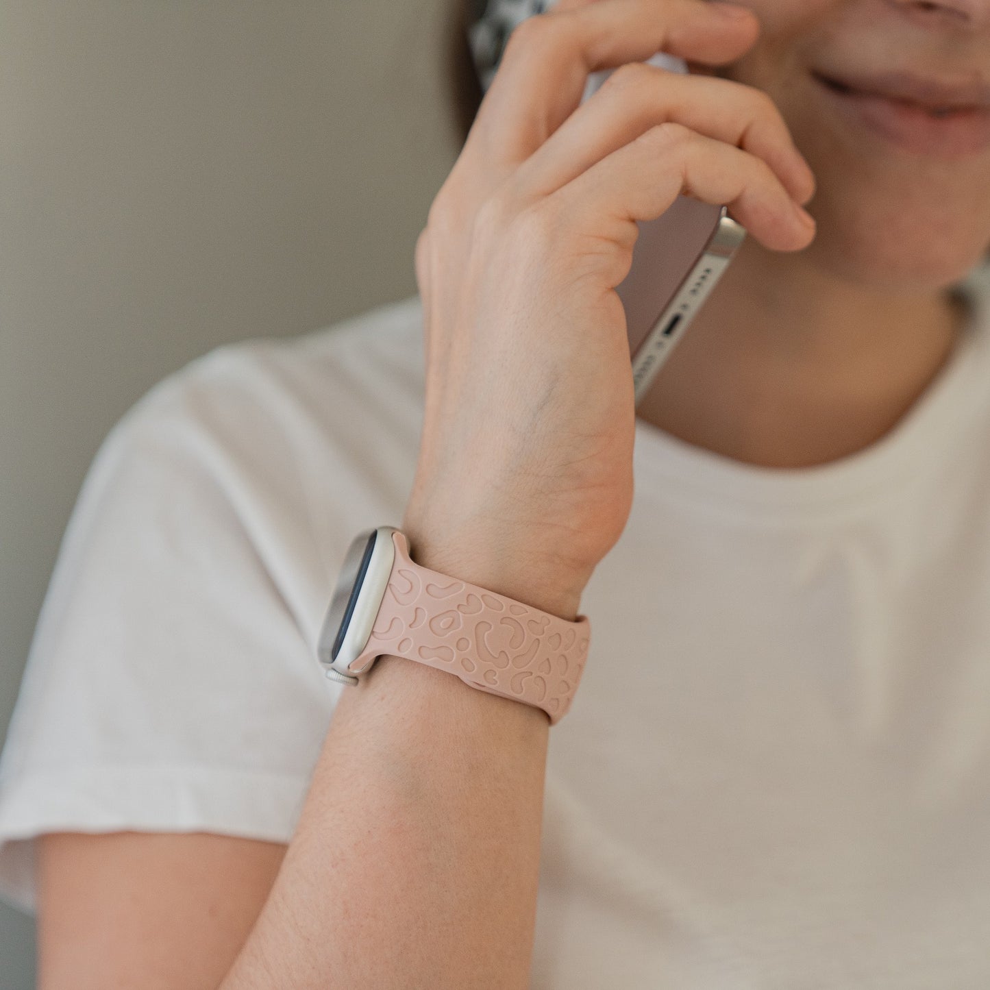 Eine Person, die ein weißes T-Shirt und ein hellrosa Arktisband-Apple-Watch-Armband „Leopard“ von Arktisband trägt, hält ein Telefon an ihr Ohr und lächelt dabei halb.