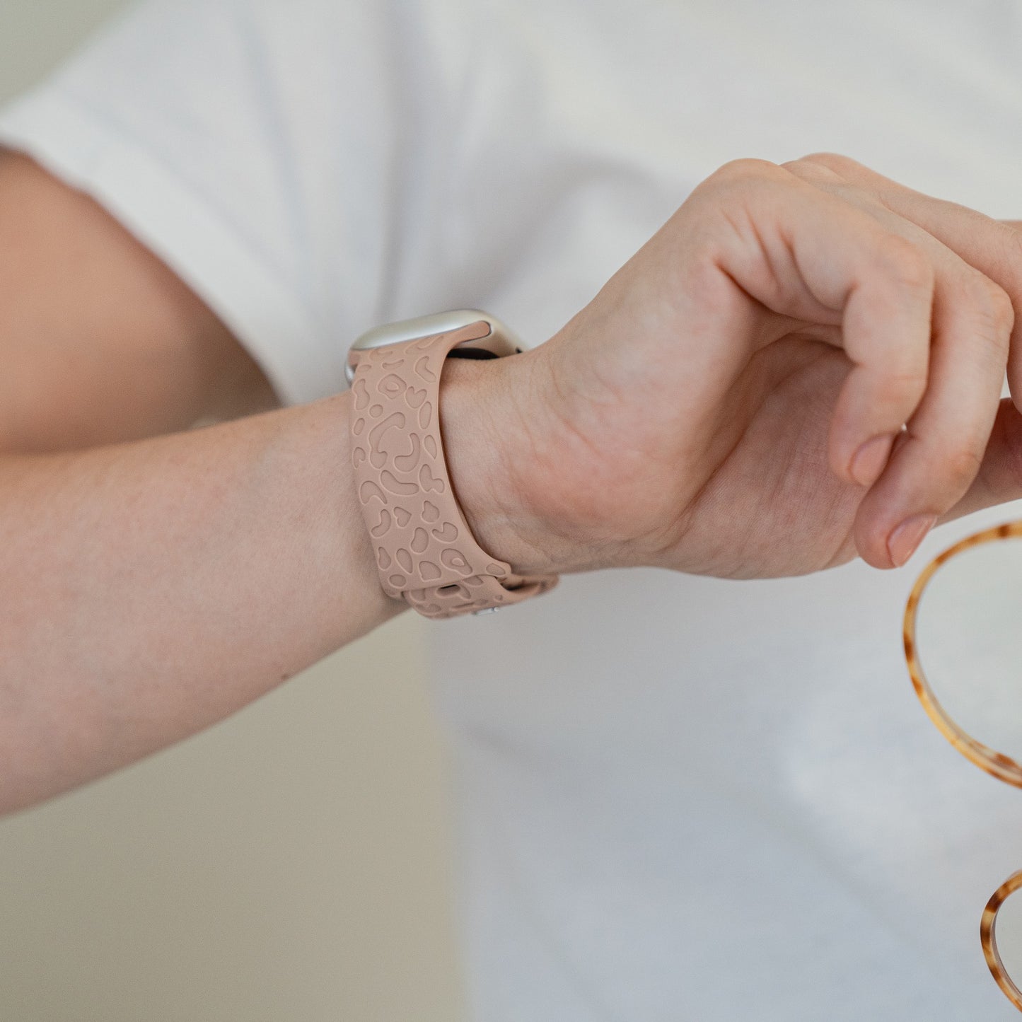 Eine Person in einem schlichten weißen Hemd rückt ihr Arktisband-Apple-Watch-Armband „Leopard“ an ihrem Handgelenk zurecht und hält in der anderen Hand einen weiteren Gegenstand mit einem braun-gelben Leopardenmuster.