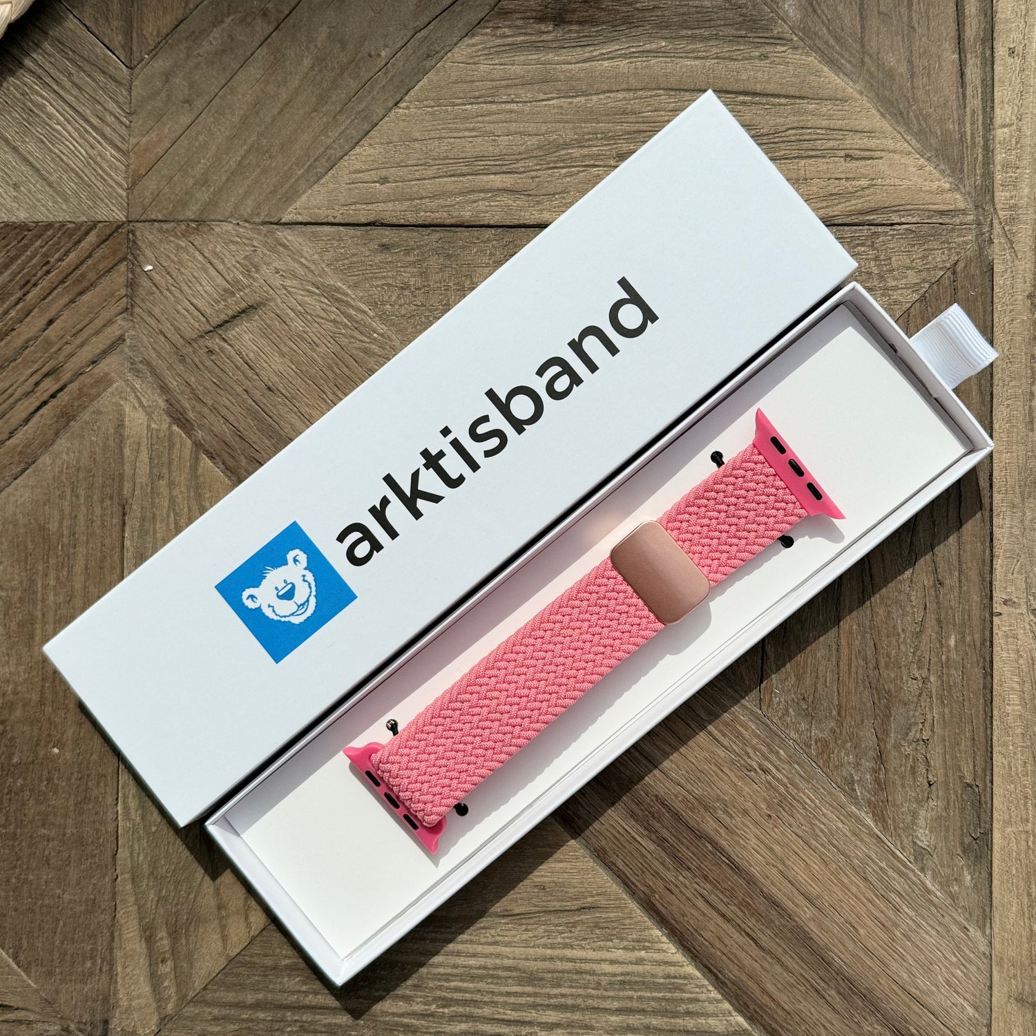 Das arktisband Geflochtene Apple Watch Armband „Casual“ mit Magnetverschluss von arktisband in Pink liegt in einer offenen weißen arktisband Box auf einer Holzoberfläche.