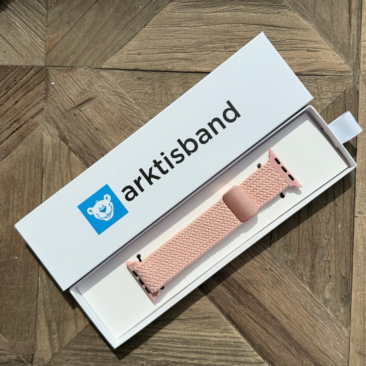 Das arktisband Geflochtene Apple Watch Armband „Casual“ mit Magnetverschluss in Pink liegt in einer offenen arktisband Box auf einer Holzoberfläche.