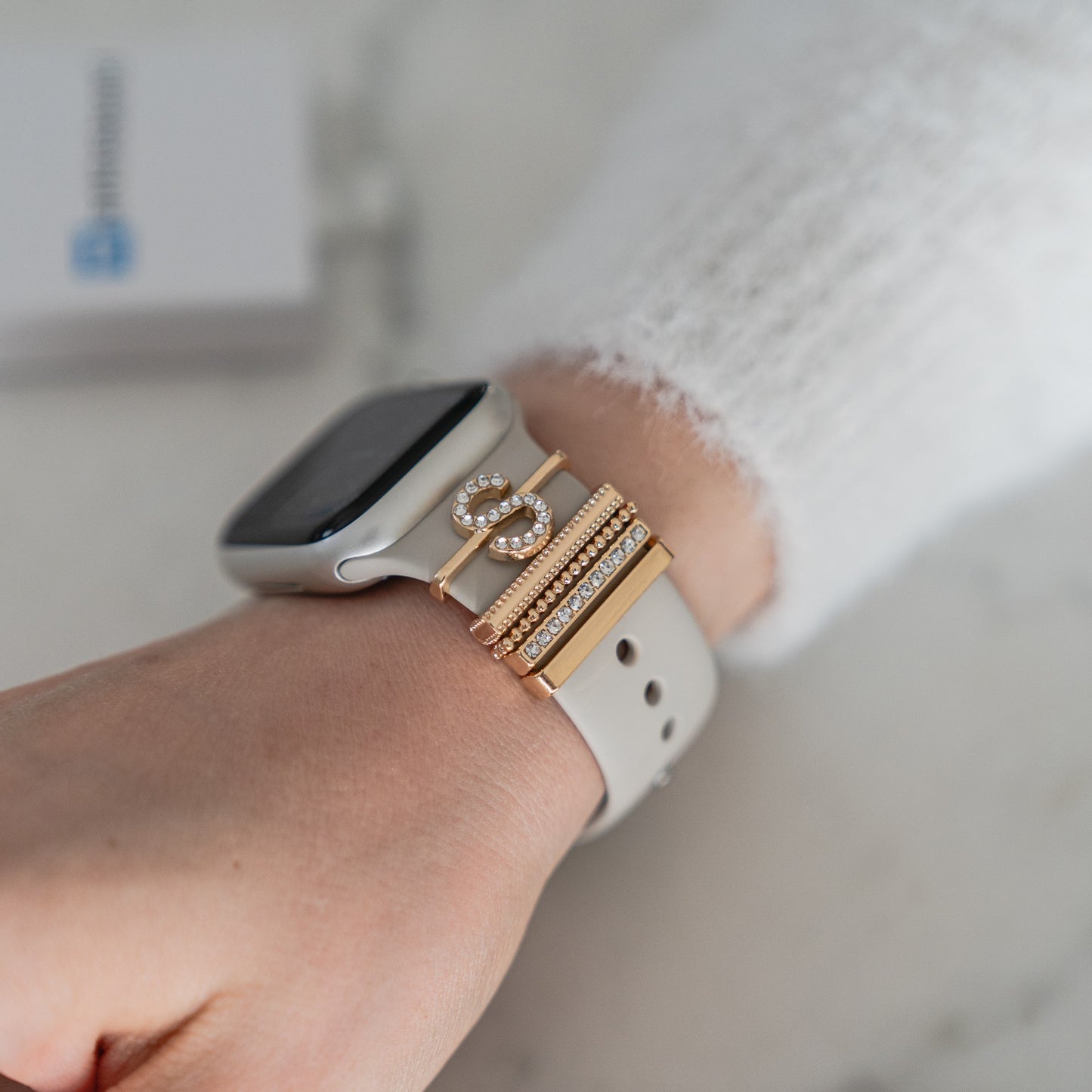 Eine Nahaufnahme eines Handgelenks mit einem hellgrauen Arktisband-Apple-Watch-Armband, verziert mit mehreren Arktisband-Apple-Watch-Charms im „S-Stil“ in Gold und Strass, gepaart mit einem weißen strukturierten Langarmshirt.