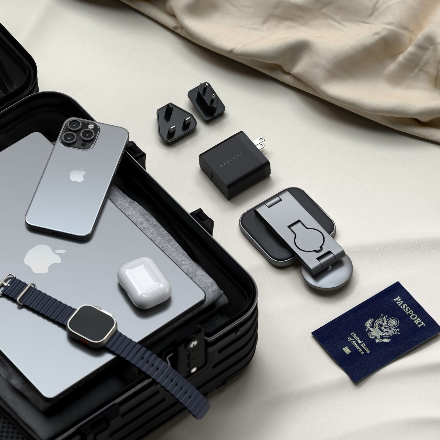 Offener Koffer auf einem Bett mit Laptop, Smartphone, Smartwatch, Kopfhörern, Adaptern, US-Reisepass und dem Satechi 3-in-1 Foldable Qi2 Wireless Charging Stand von Satechi.