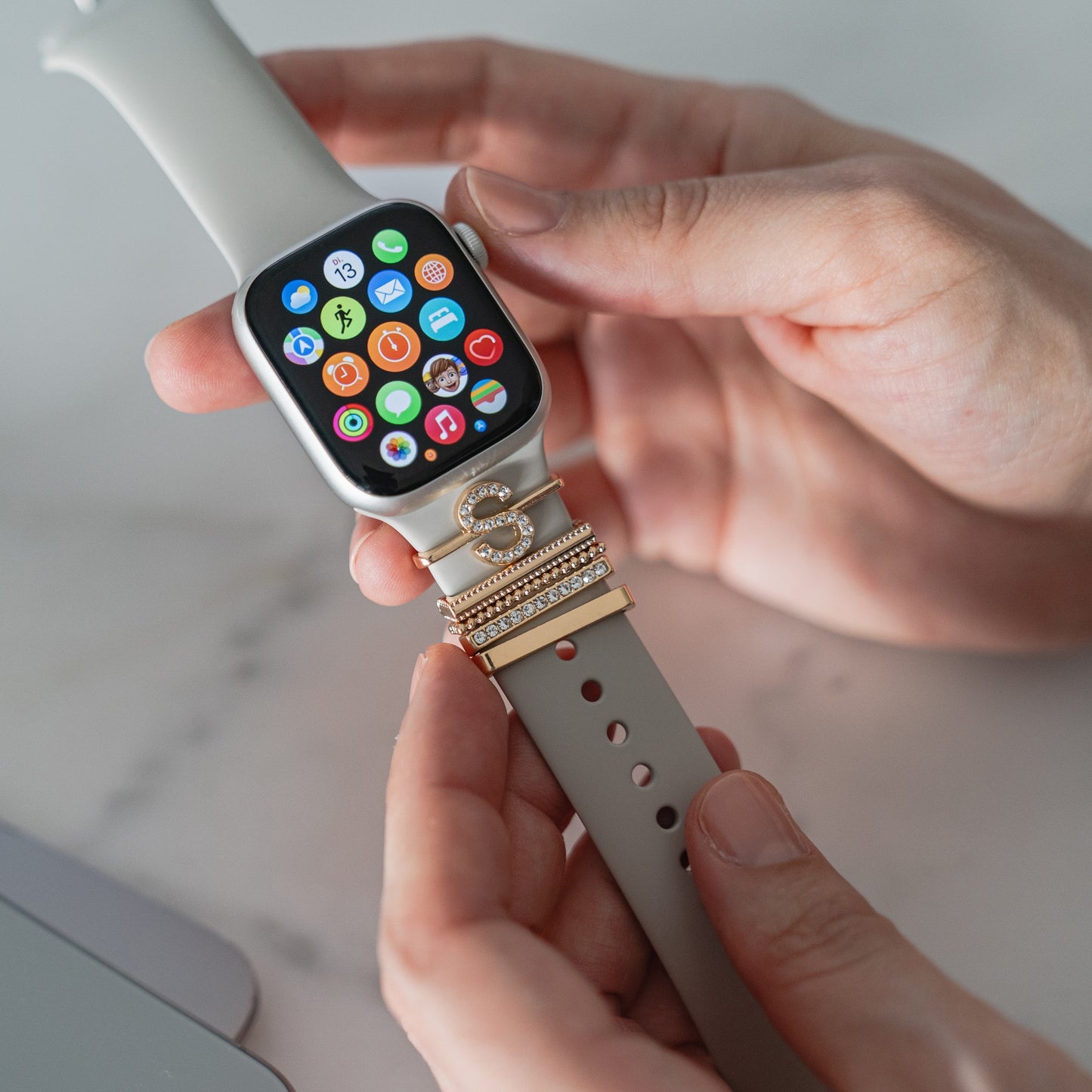 Eine Person hält ein Arktisband Apple Watch Armband, das mit Arktisband Apple Watch Charms „S Style“ verziert ist, während auf dem Bildschirm der Smartwatch mehrere bunte App-Symbole angezeigt werden.