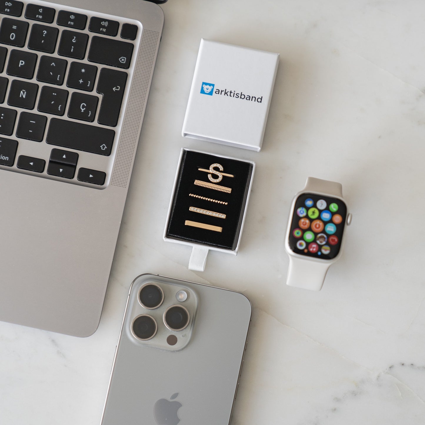 Ein MacBook, ein iPhone, eine Apple Watch mit weißem Armband und eine offene Schachtel mit goldenen Apple Watch-Charms im „S-Stil“ von Arktisband sind auf einer weißen Marmoroberfläche angeordnet.
