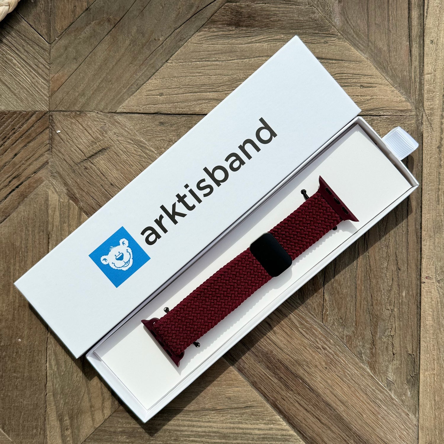 Das arktisband Geflochtene Apple Watch Armband „Casual“ mit Magnetverschluss in Maroon wird in einer offenen weißen arktisband Box auf einer Holzoberfläche präsentiert.