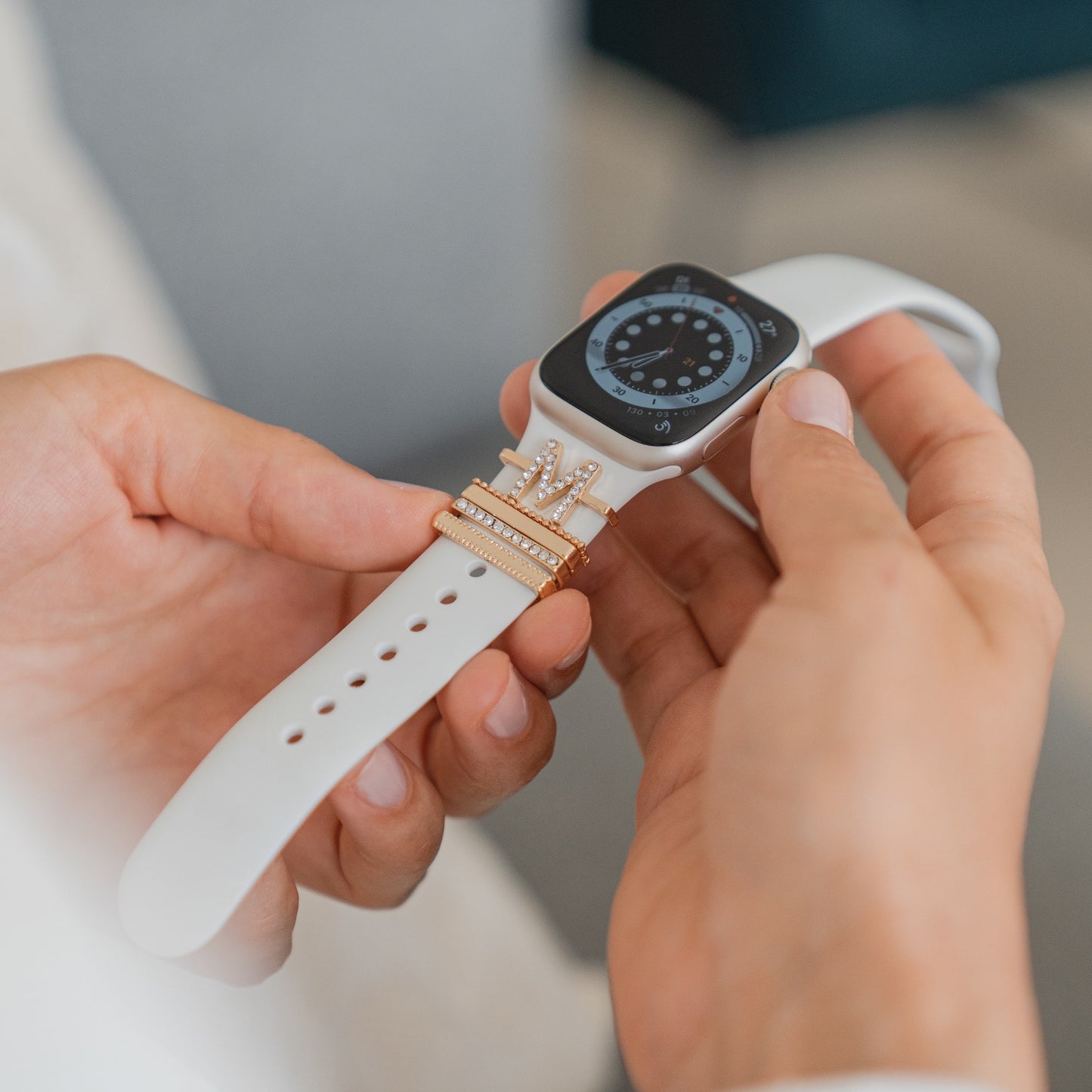 Eine Person hält eine weiße Smartwatch, verziert mit arktisband Apple Watch Charms „M Style“ mit goldenen Akzenten und Strasssteinen. Das analoge Display ergänzt das elegante Accessoire von arktisband.