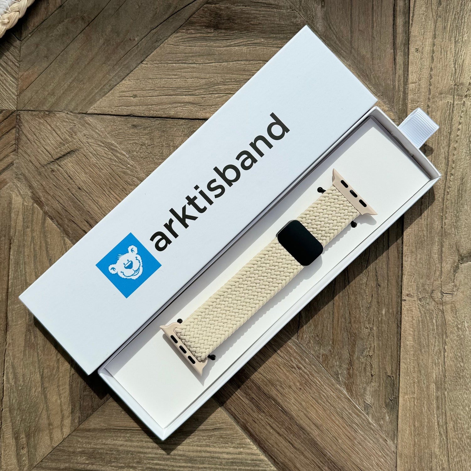 Das arktisband Geflochtene Apple Watch Armband „Casual“ mit Magnetverschluss in Creme liegt in seiner weißen arktisband-Box auf dem Holztisch, das Logo zeigt ein Bären-Gesicht im blauen Quadrat neben dem Markennamen.