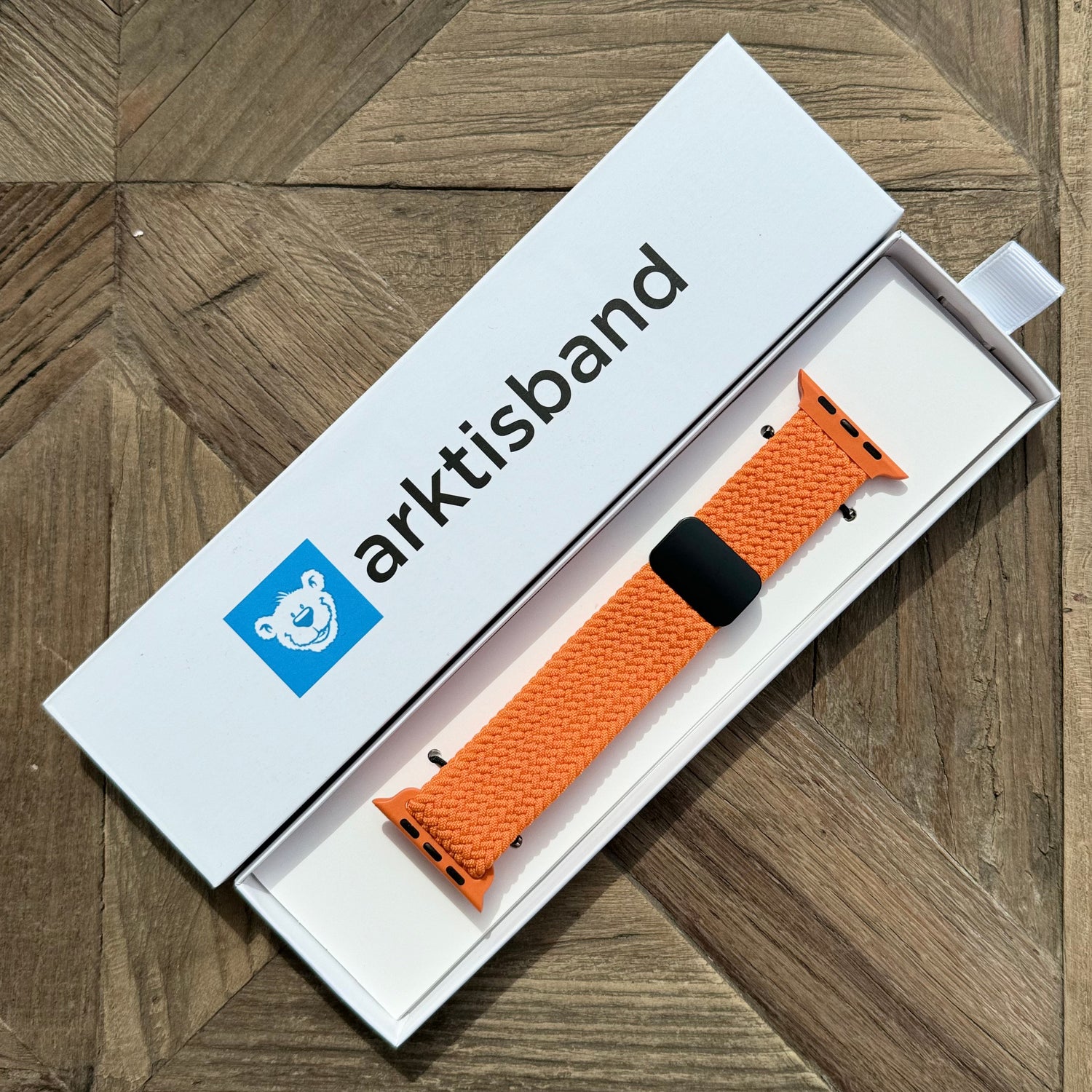 Ein orangefarbenes arktisband Geflochtenes Apple Watch Armband „Casual“ mit Magnetverschluss liegt in einer offenen weißen arktisband Box auf einer Holzoberfläche.