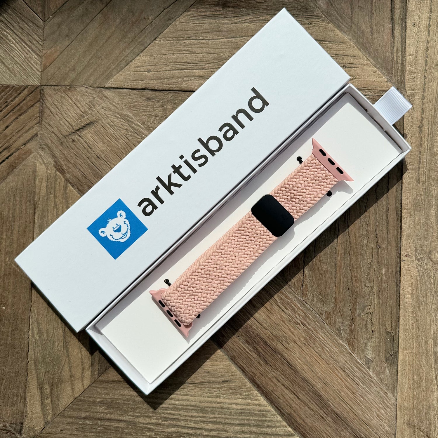 Das arktisband Geflochtene Apple Watch Armband „Casual“ mit Magnetverschluss in Pink, stilvoll präsentiert in seiner weißen Box auf einer Holzoberfläche.