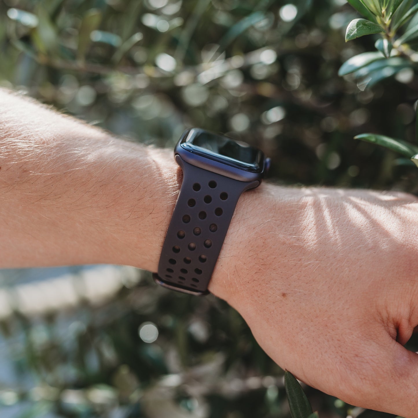 Eine Nahaufnahme des Handgelenks einer Person mit dem arktisband Apple Watch Silikonarmband „Nova“ von arktisband. Das dunkle, perforierte Silikonarmband hebt sich vor einem Hintergrund aus grünen Blättern und Sonnenlicht ab. Das Zifferblatt ist nicht sichtbar.