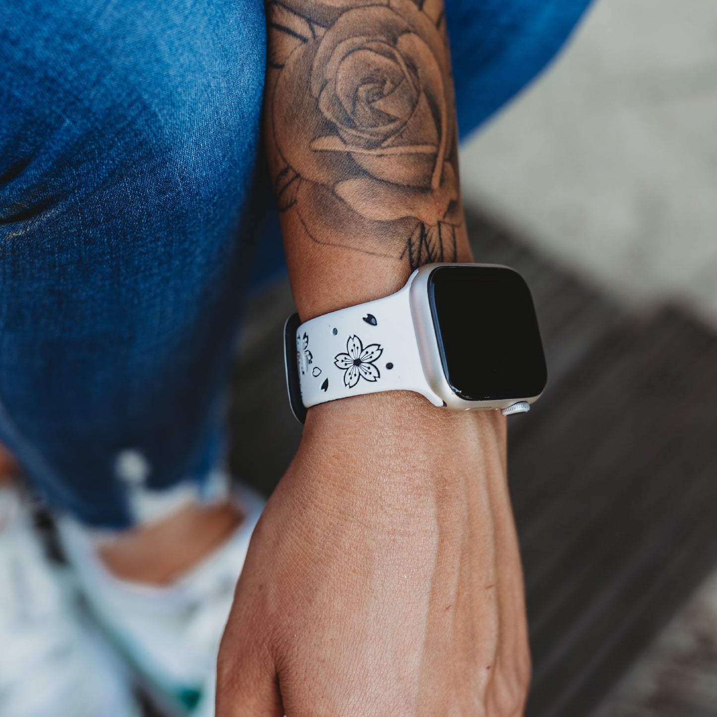 Eine Person trägt ein Arktisband Apple Watch Armband „Flores“ mit schwarzem Blumenmuster, kombiniert mit Blue Jeans, weißen Sneakers und einem Rosentattoo auf dem Unterarm.