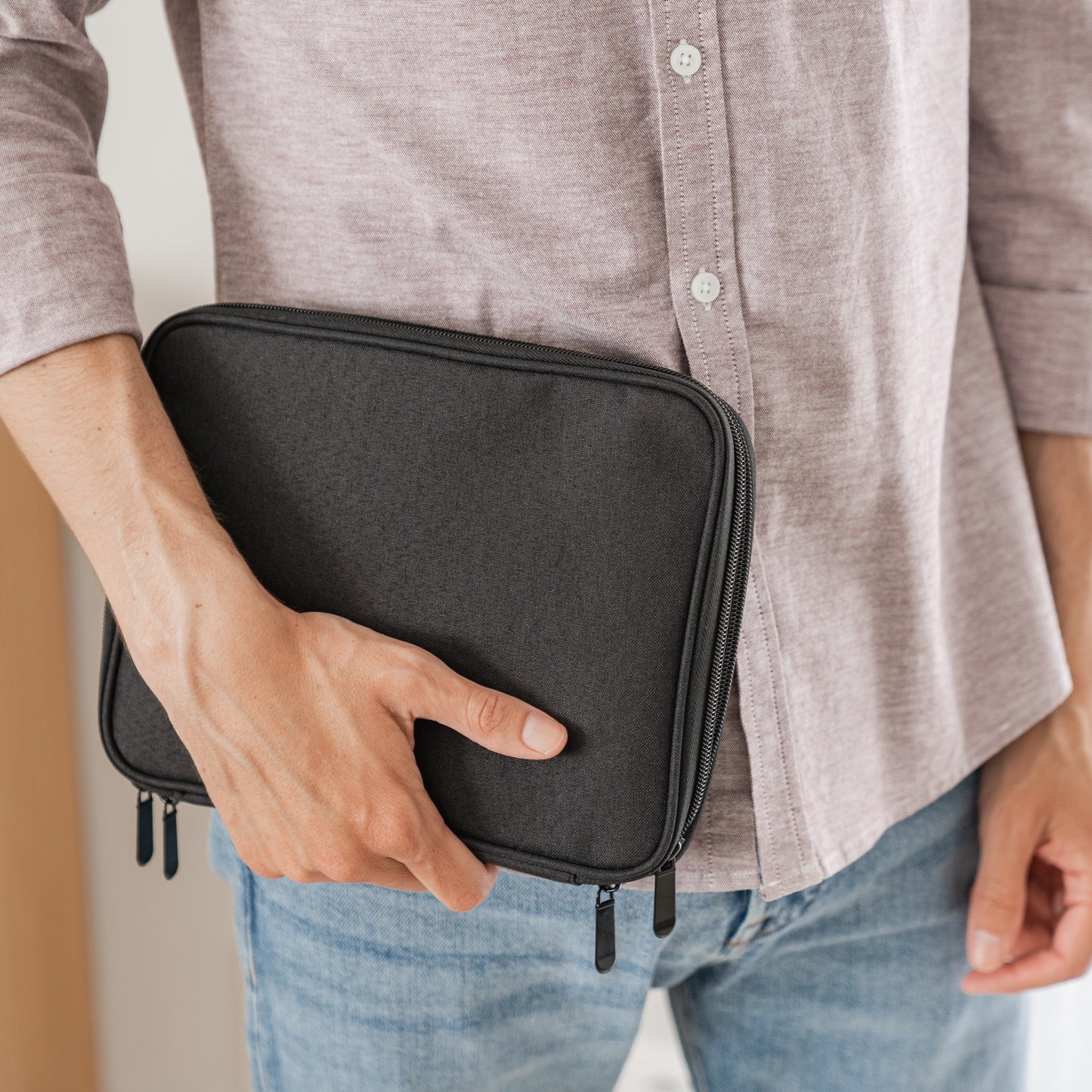 Eine Person in einem hellvioletten Hemd und blauen Jeans hält die arktisband Kleine Reisetasche für Apple Watch Armbänder & Zubehör, eine schwarze Stofftasche mit Reißverschluss von arktisband, an ihrer Seite. Es sind nur der Oberkörper und die Hand zu sehen.