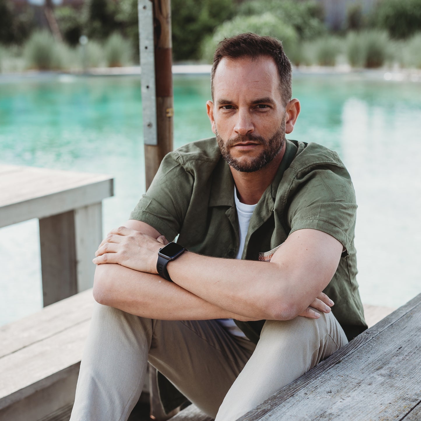 Ein Mann mit kurzen Haaren und Bart sitzt auf Holzstufen an einem Pool. Er trägt ein grünes Hemd, eine helle Hose und das arktisband Apple Watch FKM Sportarmband „Sweat“ für bequemen Alltagslook. Er stützt die Arme auf die Knie und blickt in die Kamera.