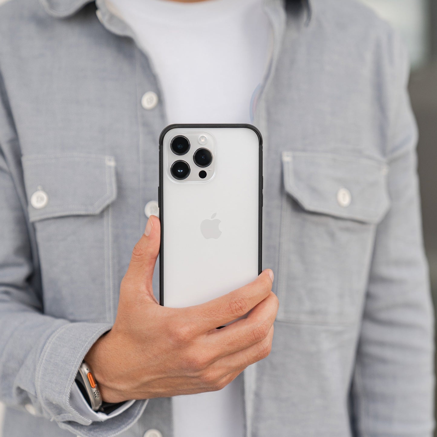 Person im grauen Hemd, die ein iPhone mit nach vorne gerichteten Linsen hält, ausgestattet mit dem Aviato AirZERO Alu Bumper für iPhone 15 Pro Max von Aviato, aus Flugzeugaluminium und ideal für kabelloses Laden.