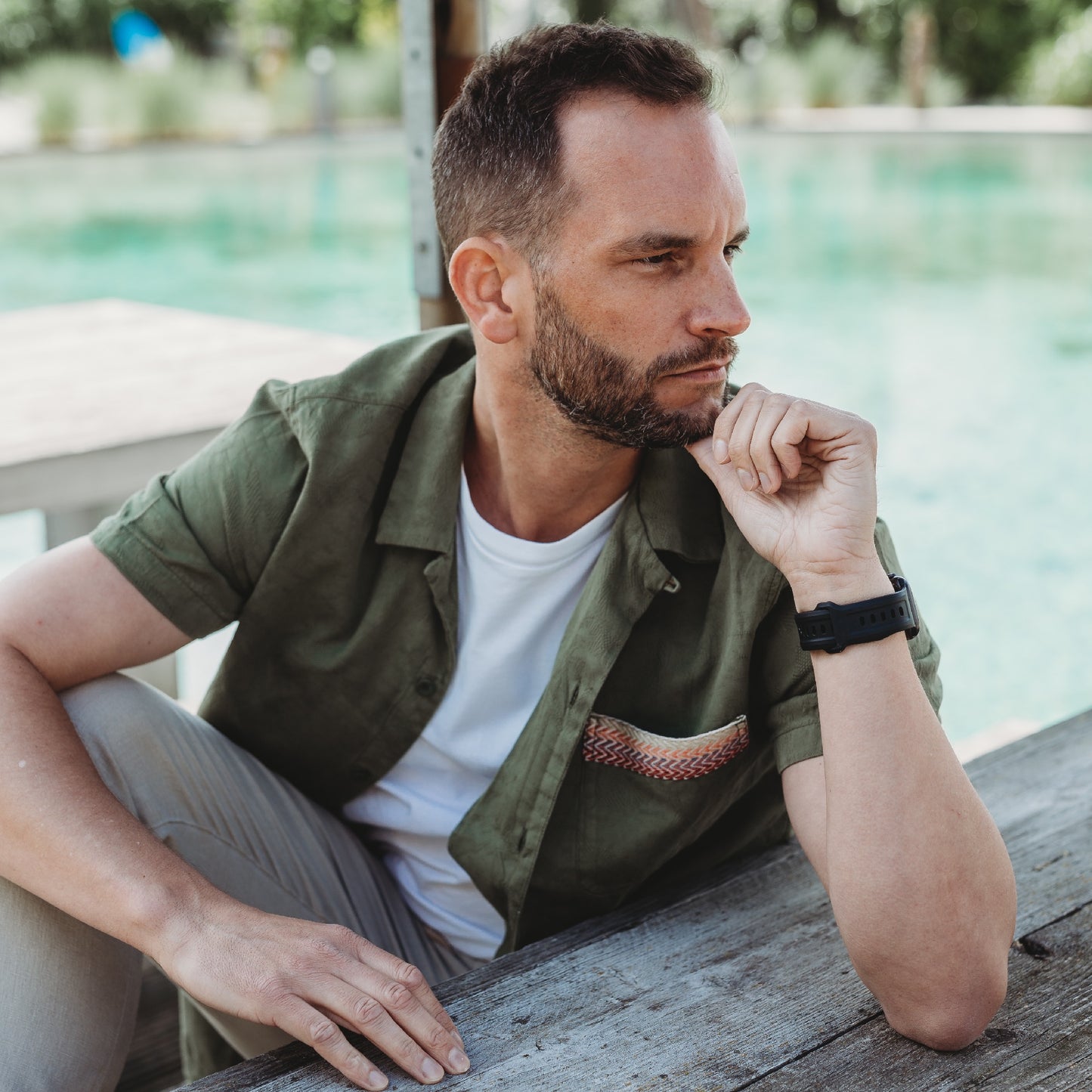 Ein Mann mit kurzen braunen Haaren und Bart sitzt an einem Pool. Er trägt ein grünes Hemd über einem weißen T-Shirt und ein Arktisband Apple Watch FKM Sportarmband „Sweat“ von Arktisband, stützt sein Kinn auf seine Hand und blickt zur Seite.