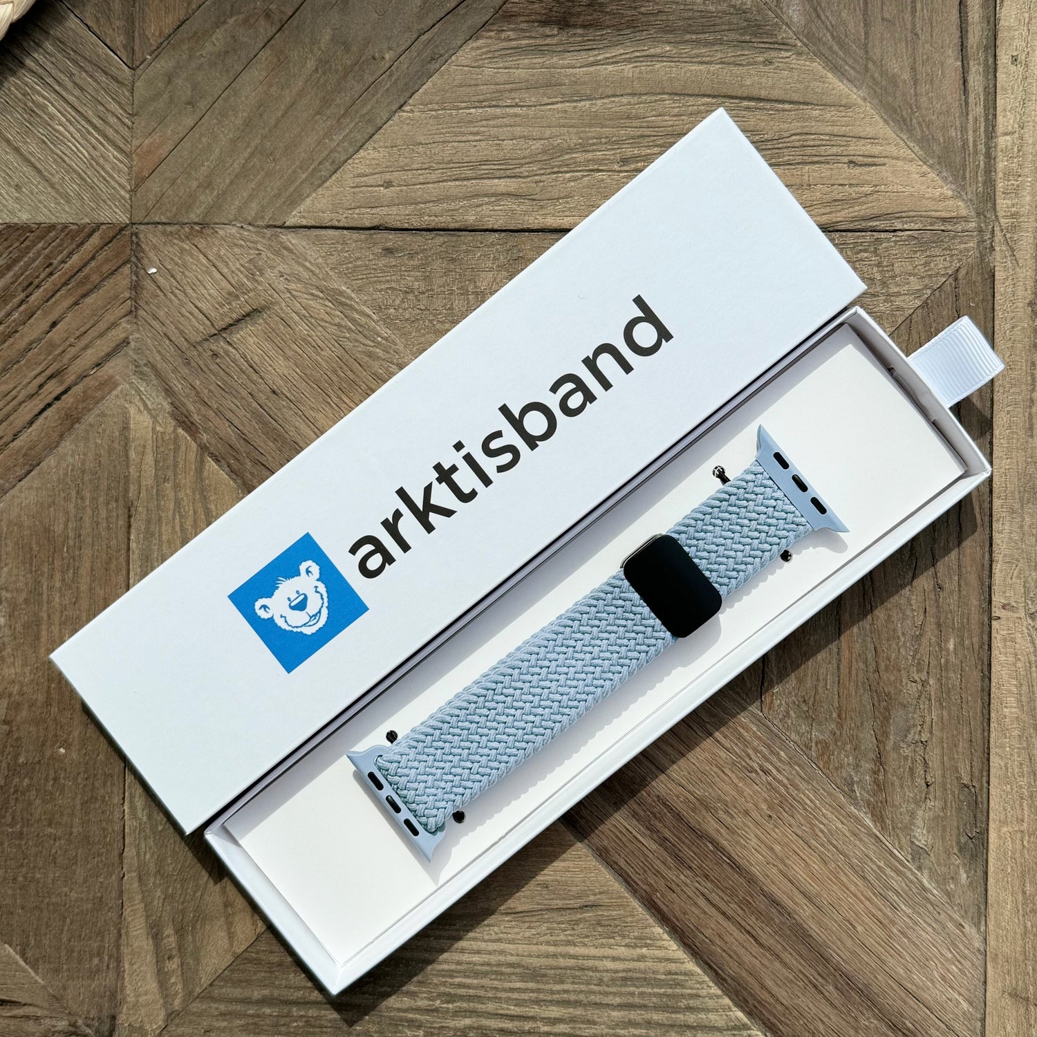 Das arktisband Geflochtene Apple Watch Armband „Casual“ mit Magnetverschluss in Hellblau wird in einer offenen weißen arktisband Box auf einer Holzoberfläche präsentiert.