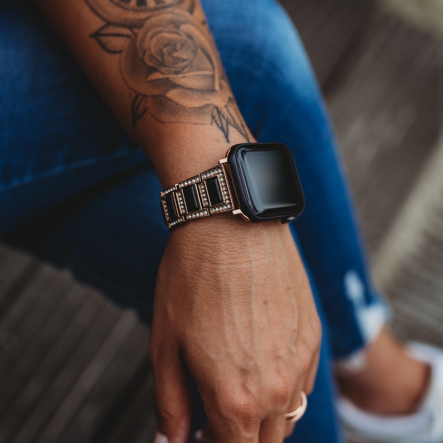 Jemand trägt ein Arktisband Apple Watch Gliederarmband „Senorita“ und sitzt in Blue Jeans auf einer Holzoberfläche und zeigt ein florales Unterarmtattoo.