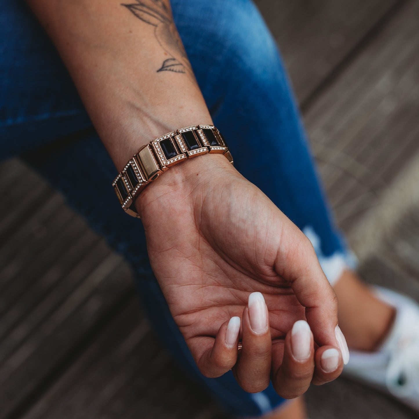 Eine Person in Bluejeans und weißen Schuhen präsentiert ihr Handgelenk mit dem arktisband Apple Watch Gliederarmband „Senorita“ von arktisband in Gold und Schwarz, ergänzt durch manikürte Nägel und ein florales Unterarmtattoo.