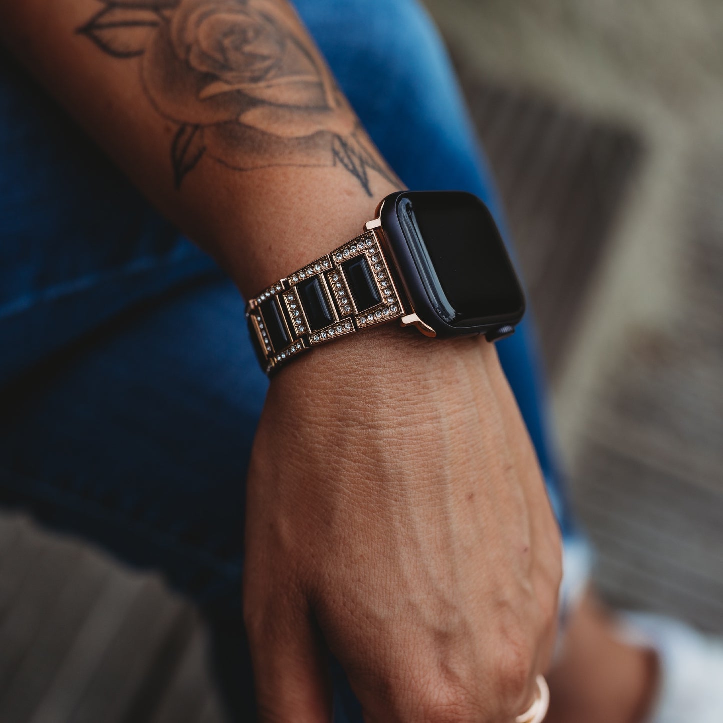 Eine Person mit Rosentattoo trägt das arktisband Apple Watch Gliederarmband „Senorita“ von arktisband, während ihre Hand auf ihrem Bein in Blue Jeans ruht.