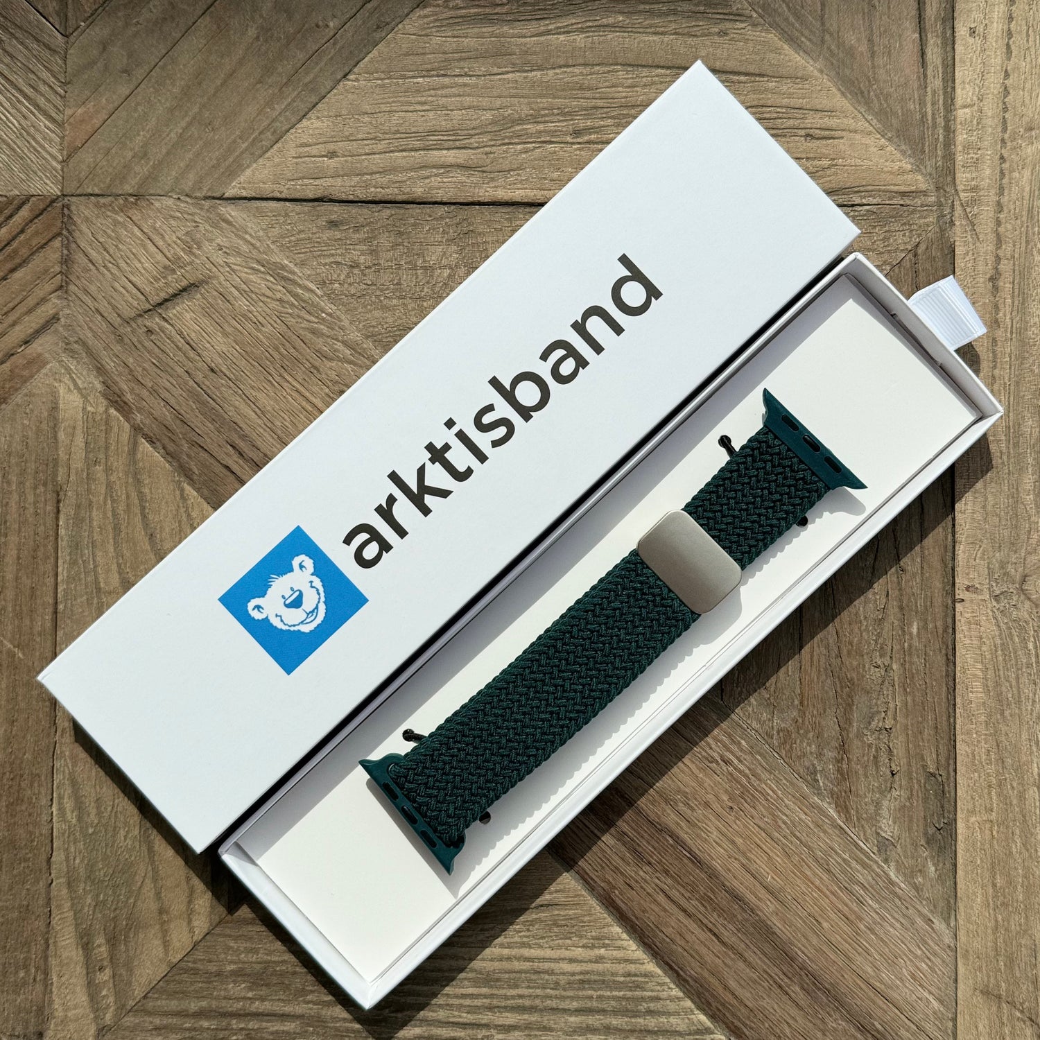 Ein dunkelgrünes arktisband Geflochtenes Apple Watch Armband „Casual“ mit Magnetverschluss liegt in einer weißen arktisband Box auf einer Holzoberfläche.