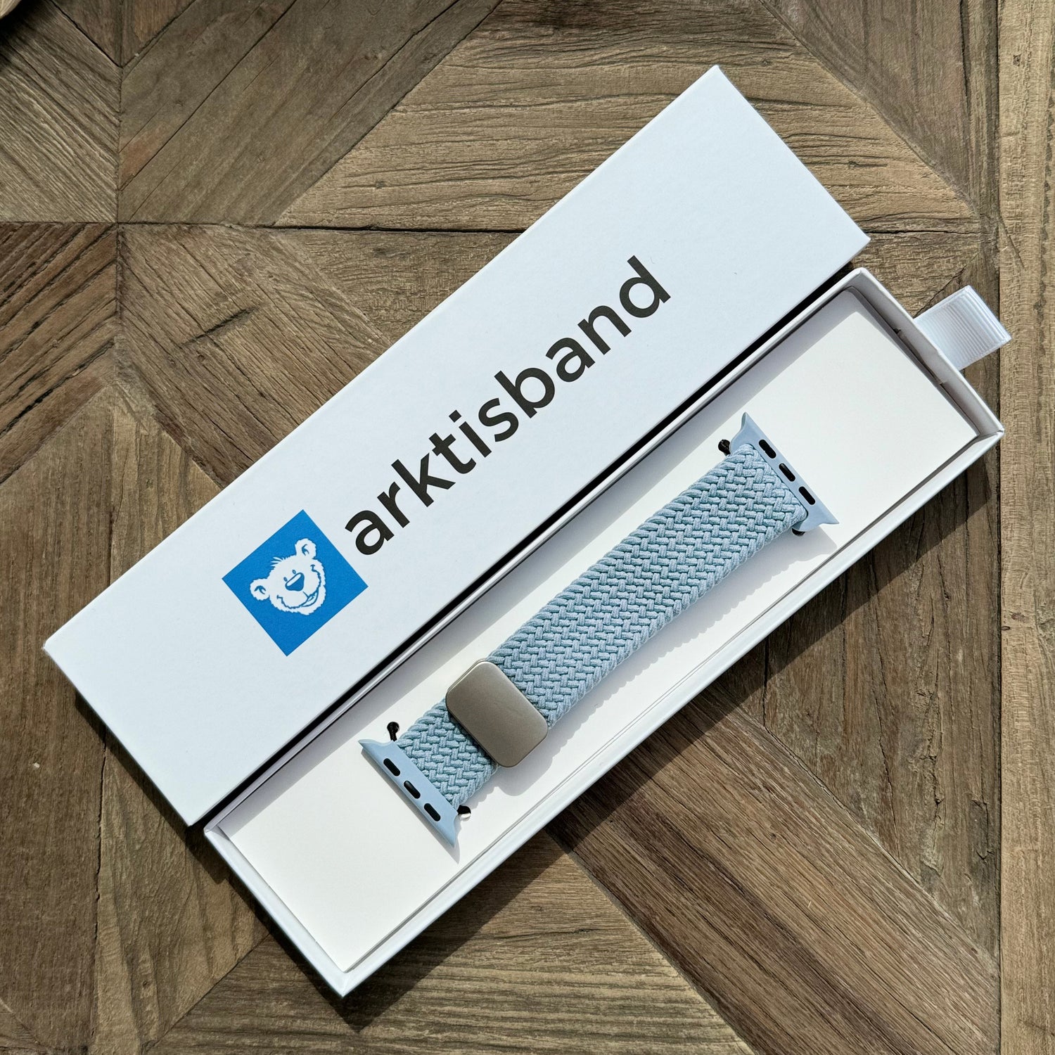 Das arktisband Geflochtene Apple Watch Armband „Casual“ in Hellblau mit Magnetverschluss von arktisband, elegant präsentiert in einer offenen weißen Box auf einer Holzoberfläche.