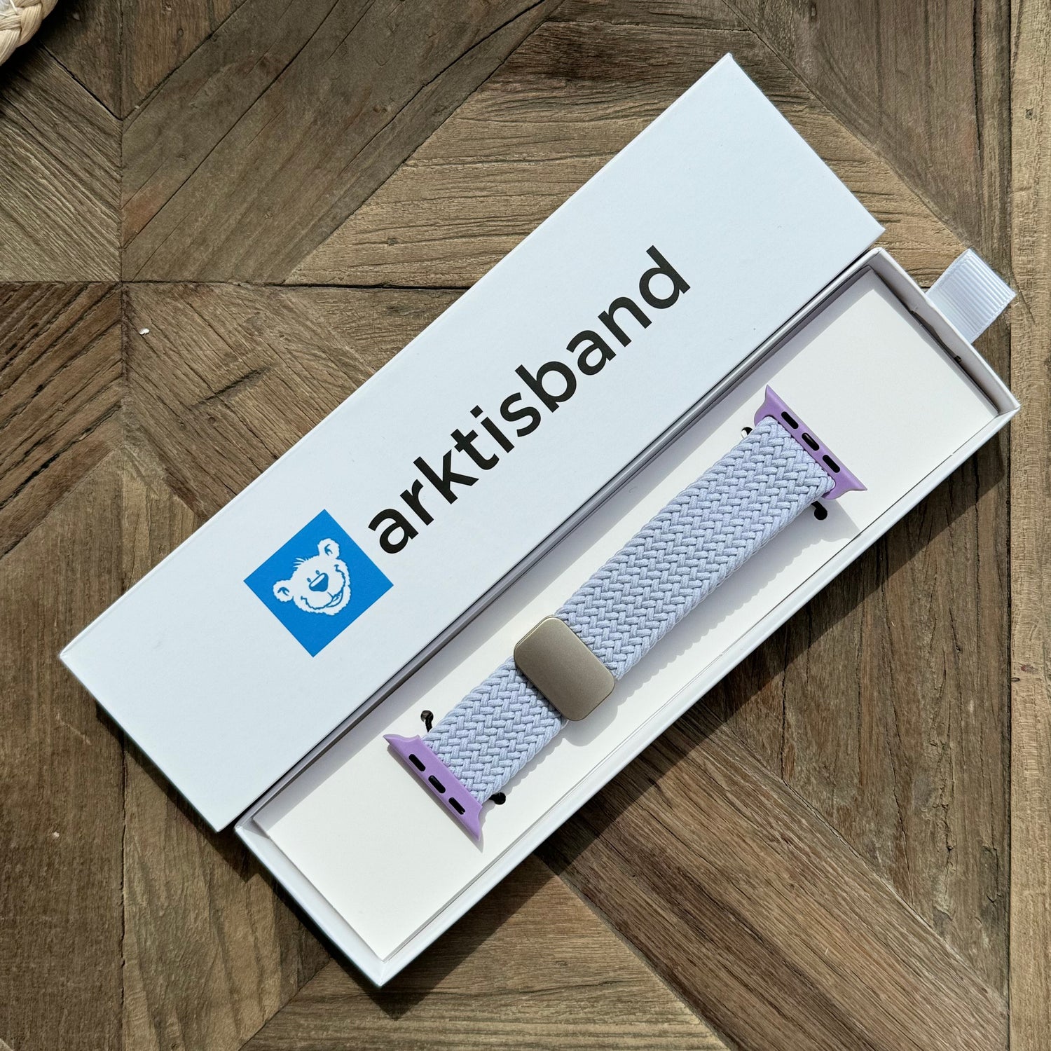 Ein verpacktes arktisband Geflochtenes Apple Watch Armband „Casual“ mit Magnetverschluss von arktisband, mit lavendelfarbenen Anschlüssen, ruht auf einer Holzoberfläche.