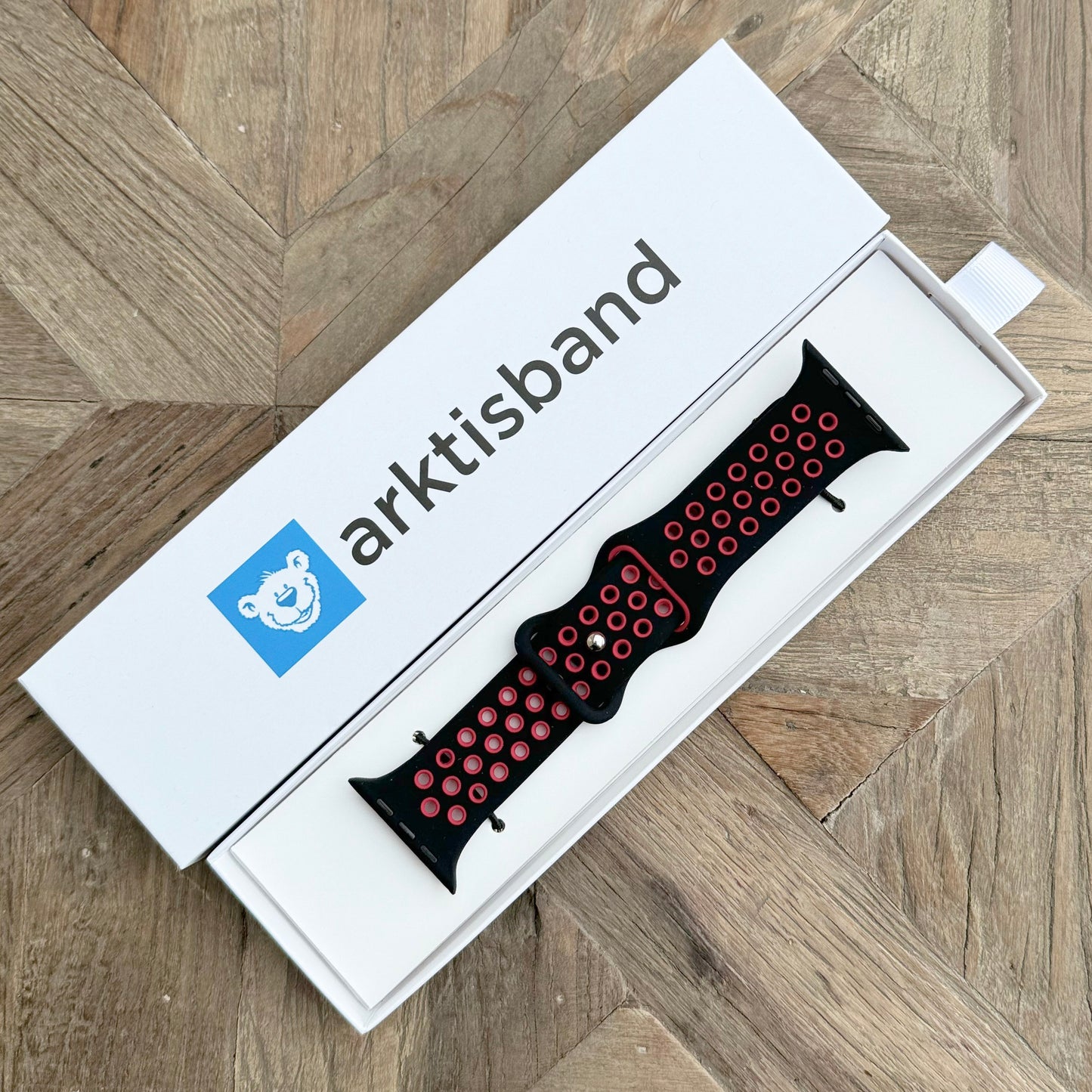 Das arktisband Apple Watch Silikonarmband „Energy“ in Schwarz mit roten Akzentlöchern wird in einer offenen weißen Box mit blauem arktisband-Bärenlogo auf einer Holzoberfläche präsentiert.