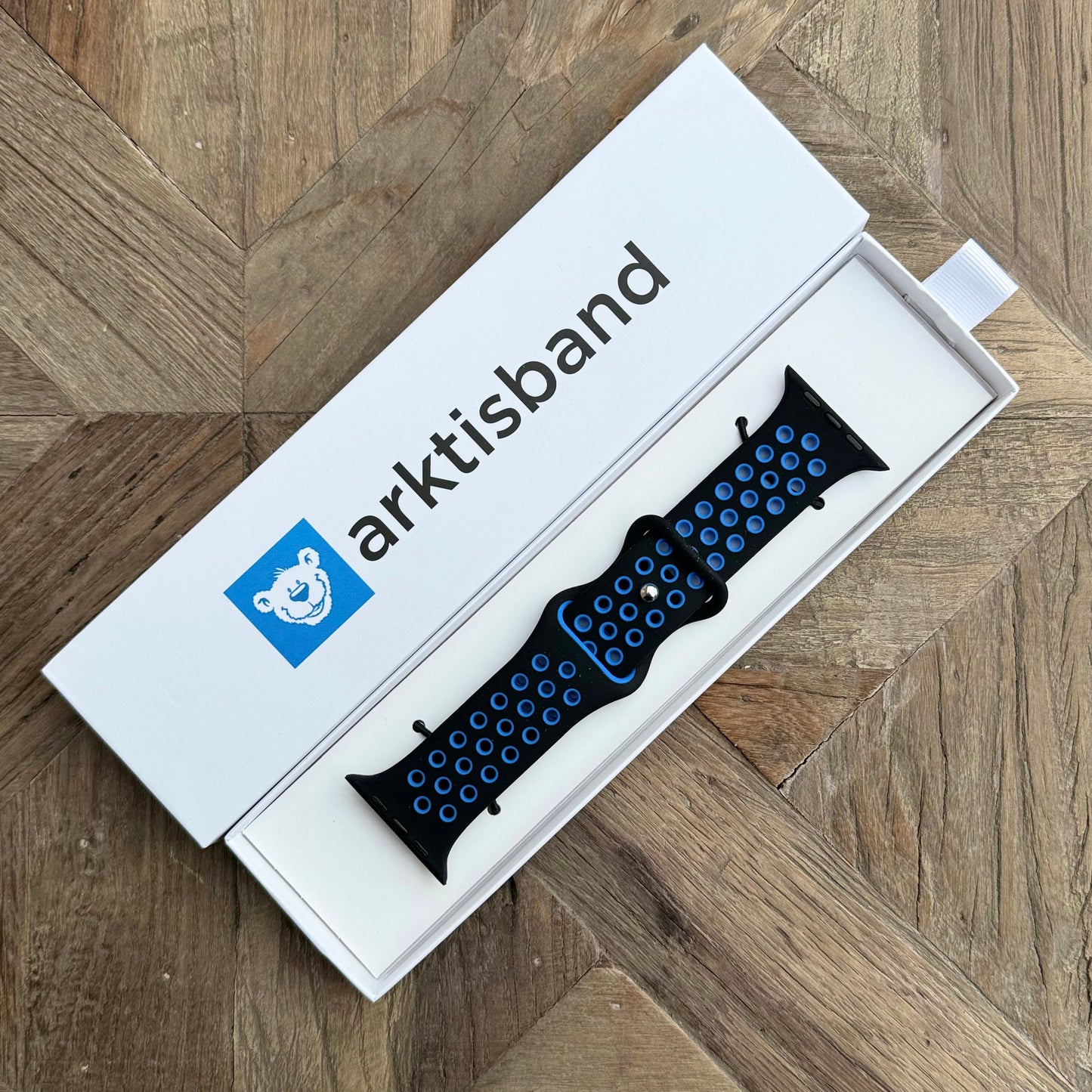 Das arktisband Apple Watch Silikonarmband „Energy“ in Blau und Schwarz wird in einer weißen Box mit dem arktisband-Namen und dem Eisbär-Logo auf dem Deckel gezeigt und ruht auf einer Holzoberfläche mit einem geometrischen Muster.