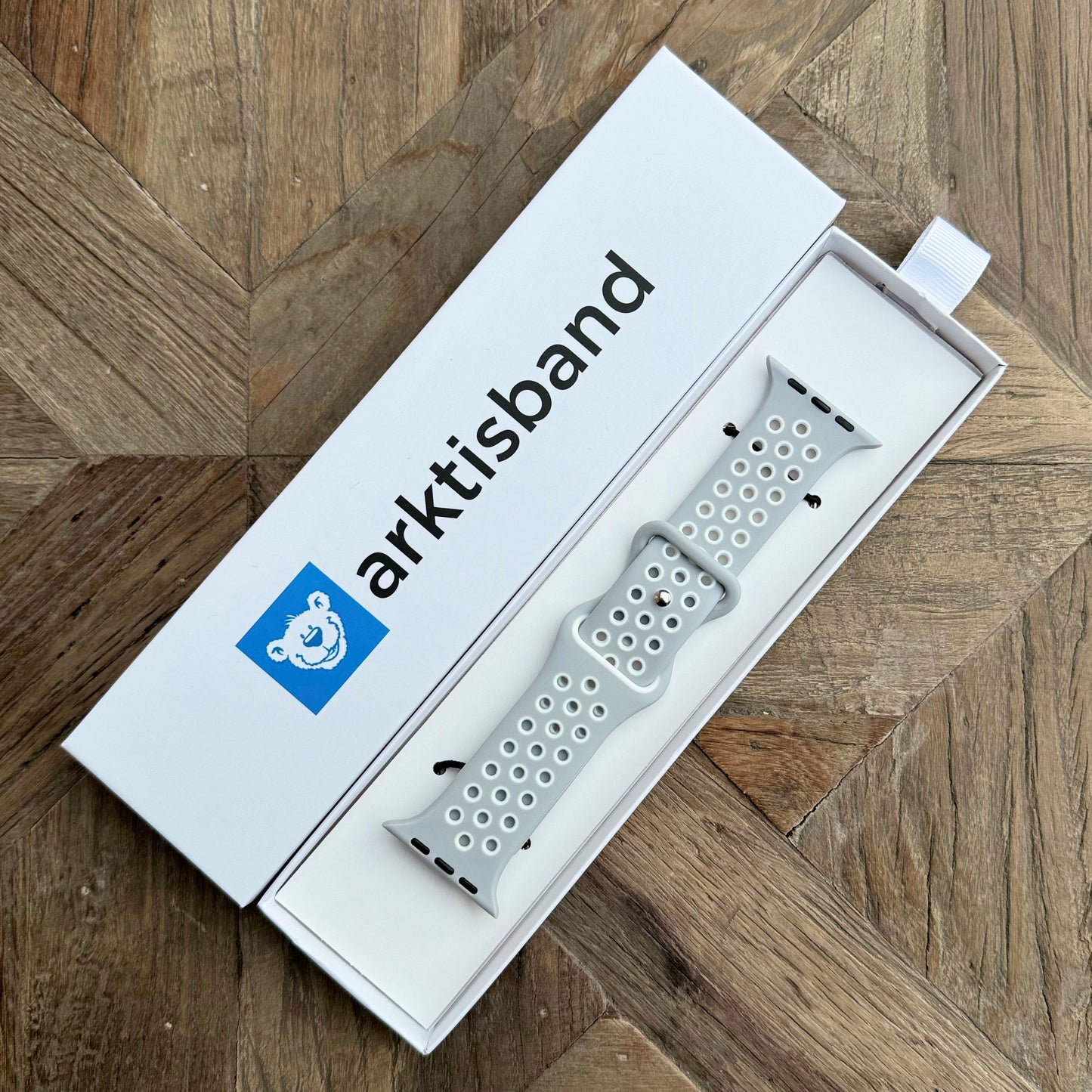 Das arktisband Apple Watch Silikonarmband „Energy“ in Hellgrau wird in einer weißen arktisband-Box mit blauem Bärenlogo gezeigt, die auf einer Holzoberfläche mit Fischgrätenmuster ruht.