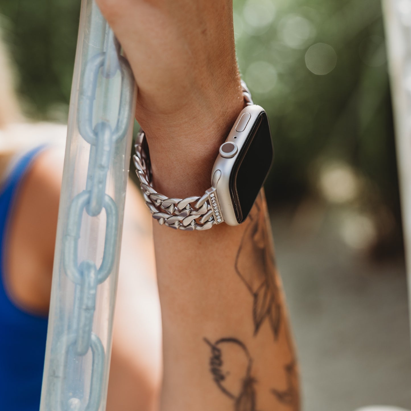 Ein tätowierter Arm greift nach einer durchsichtigen Kette, während er das arktisband Apple Watch Armband „Princess“ von arktisband trägt, dessen glänzende silberne Glieder einen Kontrast zu einem verschwommenen grünen Hintergrund bilden.