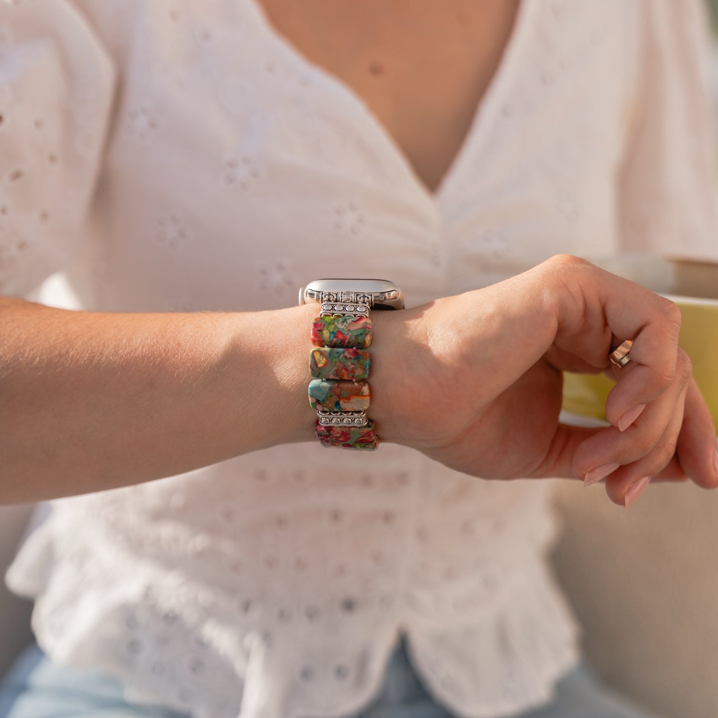 Eine Frau in einer weißen Bluse hält eine gelbe Tasse und trägt die arktisband Apple Watch „Stone Loop“ von arktisband, ein farbenfrohes und möglicherweise mit Naturstein-Akzenten versehenes Armband.