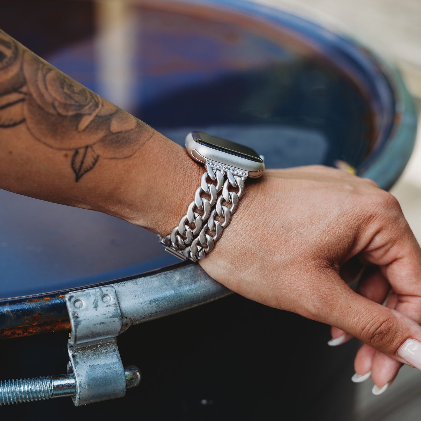 Eine Person mit einem floralen Unterarmtattoo legt ihre Hand auf eine blaue Metalltrommel und trägt das arktisband Apple Watch Armband „Princess“ von arktisband, das eine glanzvolle Gliederstruktur und ein auffälliges Kettenglieddesign aufweist.