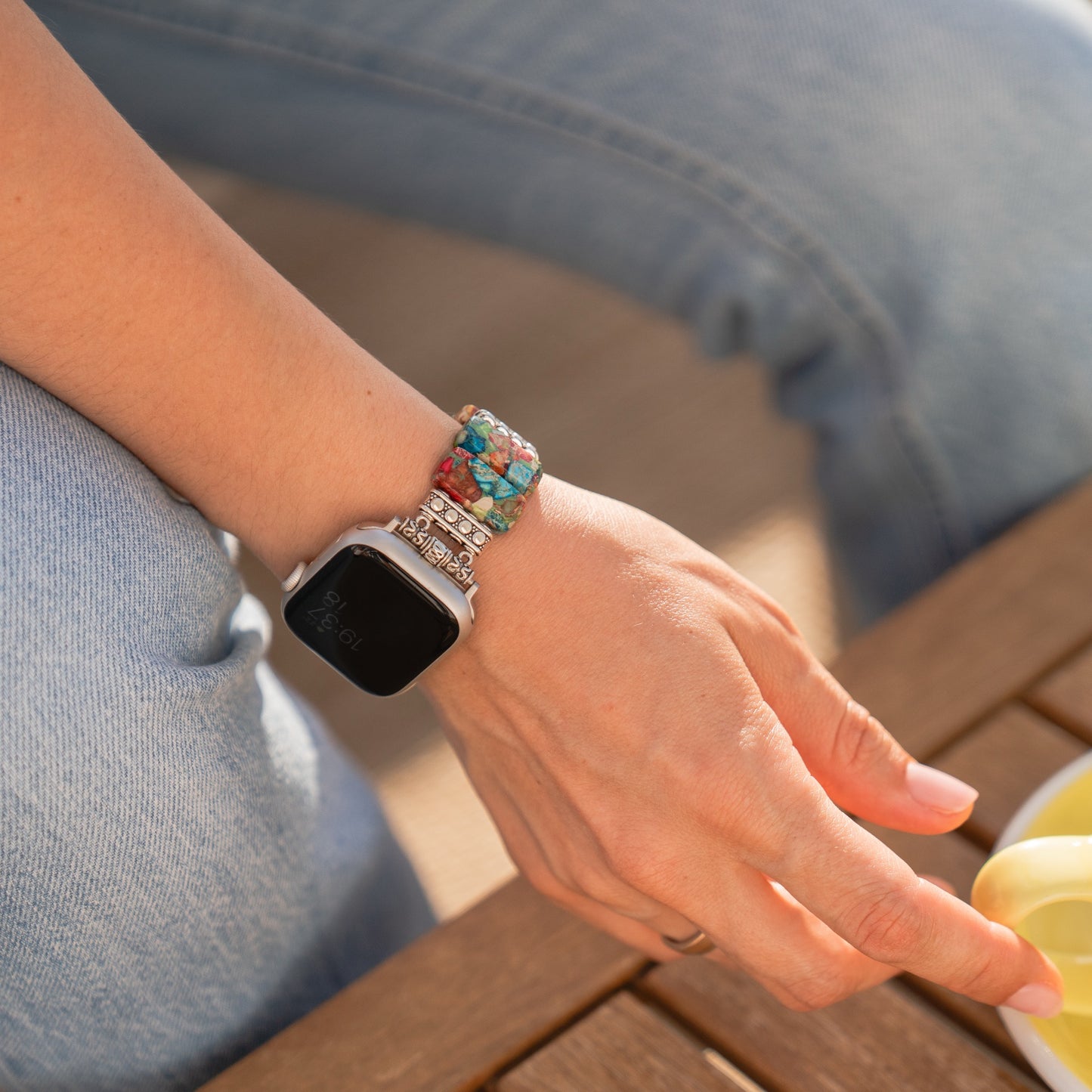 Jemand trägt ein Arktisband Apple Watch „Stone Loop“-Armband mit bunten Perlen und Natursteinen und sitzt im Sonnenlicht an einem Holztisch.