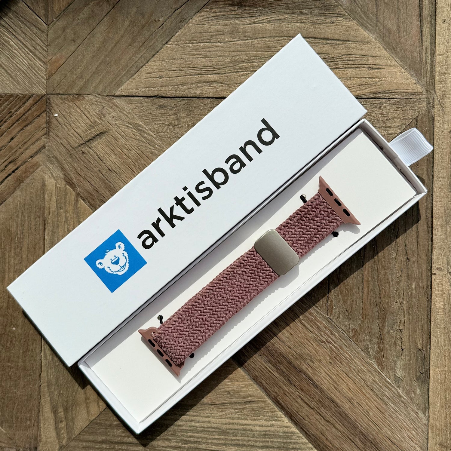Das arktisband Geflochtene Apple Watch Armband „Casual“ mit Magnetverschluss in Pink wird in einer offenen weißen arktisband Box auf einer Holzoberfläche präsentiert.