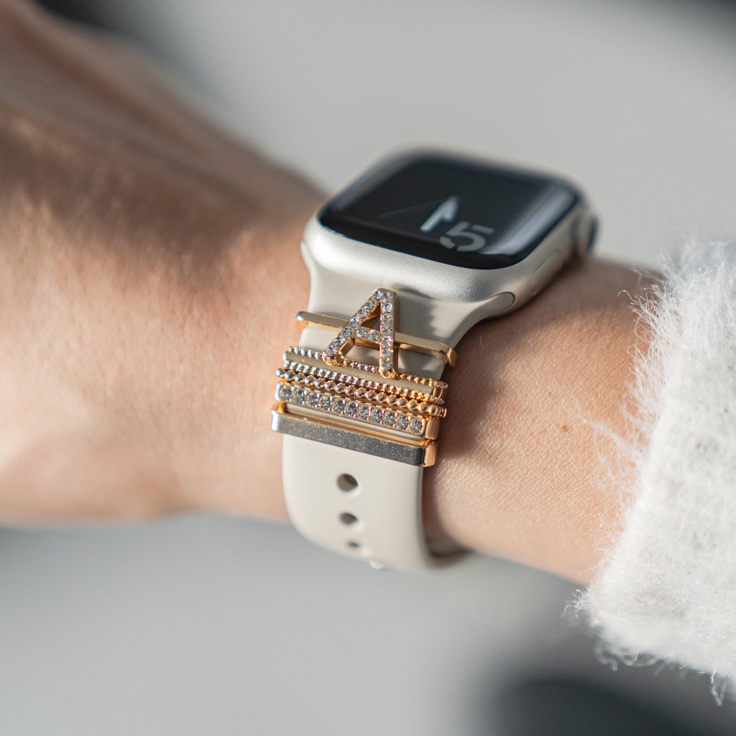 Ein Arktisband Apple Watch Charms „A Style“ – gestapelte A-Charms aus Gold und Kristall – schmücken ein beiges Band am Handgelenk.