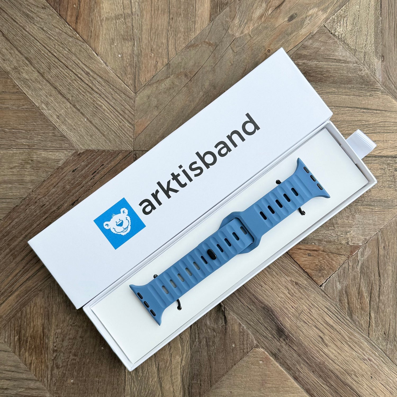 Ein arktisband Apple Watch Silikonarmband „Active“ in Blau, verpackt in einer weißen Box mit dem arktisband-Logo, liegt auf einer hölzernen Fischgrätenoberfläche und betont seinen atmungsaktiven Komfort und sein modernes Design.