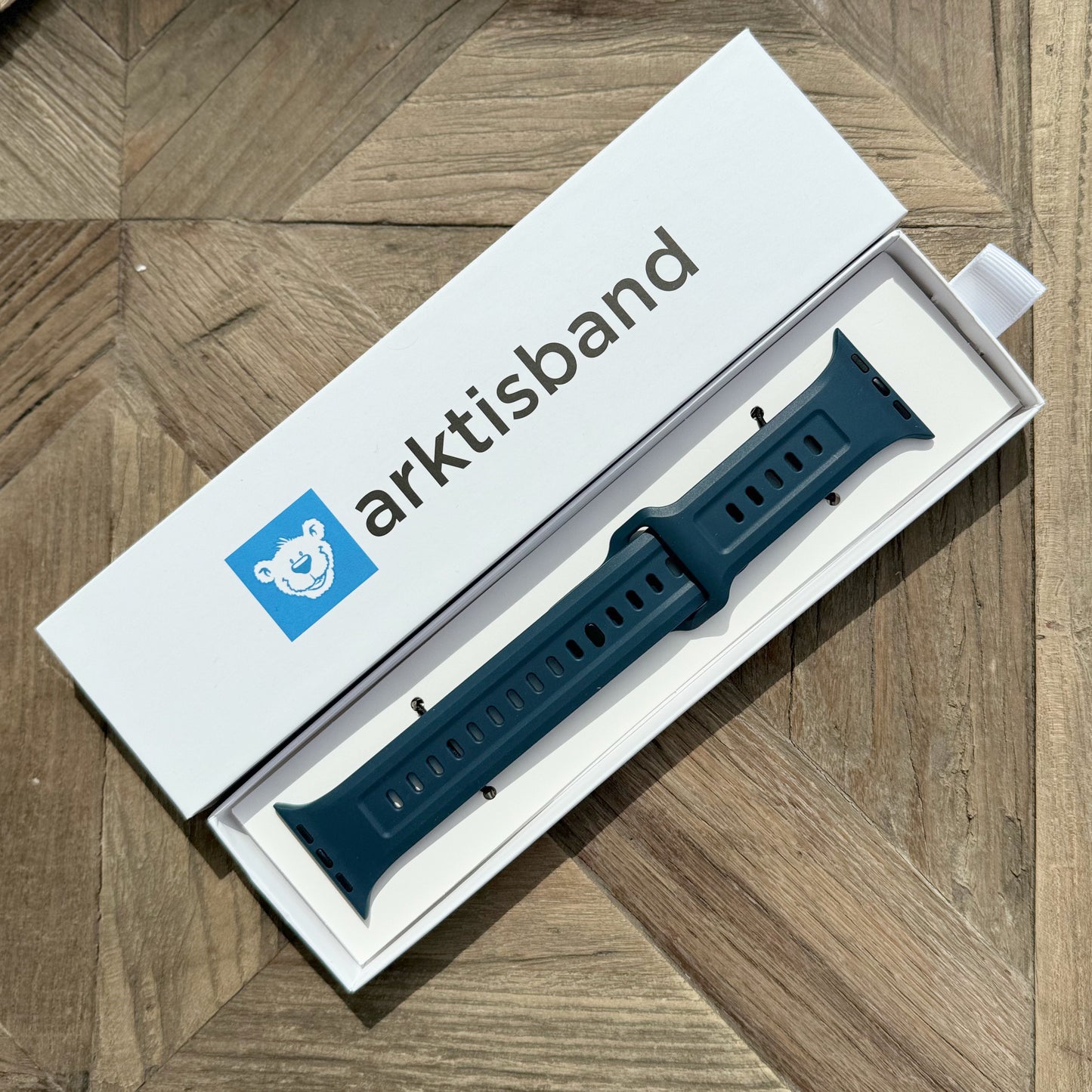 Das arktisband Apple Watch FKM Sportarmband "Sweat" aus dunkelblaugrünem Fluorelastomer wird in einer weißen arktisband-Box mit einem blauen Bärenlogo auf gemustertem Holz geliefert. Strapazierfähig, elastisch und Apple Watch kompatibel.