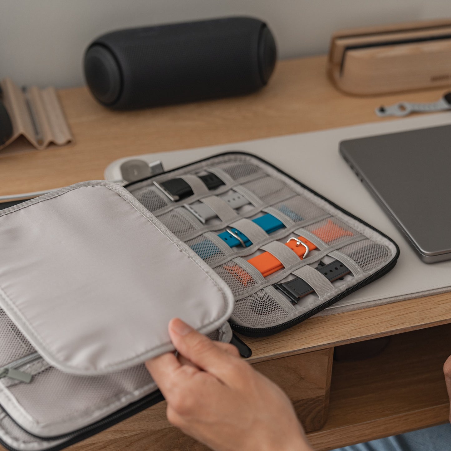 Eine Person öffnet die arktisband Kleine Reisetasche für Apple Watch Armbänder & Zubehör und enthüllt darin bunte Armbänder. Der graue Organizer von arktisband steht auf einem Holzschreibtisch, im Hintergrund ein Laptop, ein Lautsprecher und Zubehör.