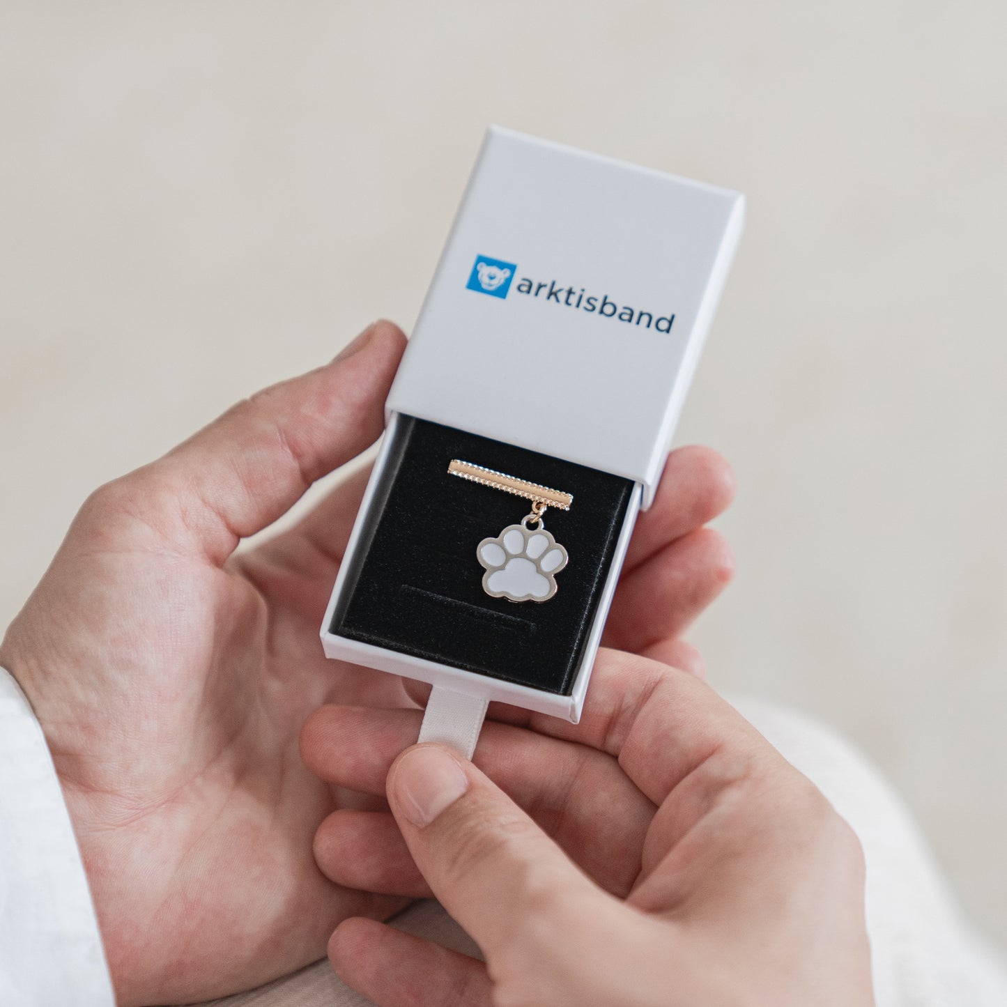 Eine Person hält eine offene weiße Arktisband-Box, aus der eine goldene Halskette mit Pfotenanhänger auf einem schwarzen Kissen hervorgeht – perfekt passend zu den Arktisband-Armbandanhängern für Ihre Apple Watch.