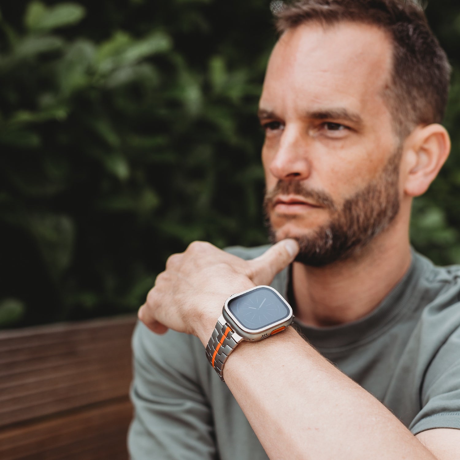 Ein Mann mit gestutztem Bart und grünem Hemd sitzt im Freien auf einer Bank, stützt sein Kinn auf seine Hand und präsentiert das Arktisband Apple Watch TITAN Armband „Aviato Edition“. Im Hintergrund ist grünes Laub zu sehen.