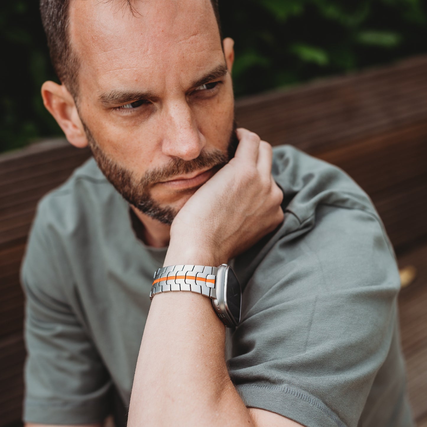 Ein Mann mit gestutztem Bart und grünem T-Shirt sitzt auf einer Holzbank und stützt sein Kinn auf seine Hand. An seinem linken Handgelenk trägt er das arktisband Apple Watch TITAN Armband „Aviato Edition“. Grünes Laub bildet den Hintergrund.