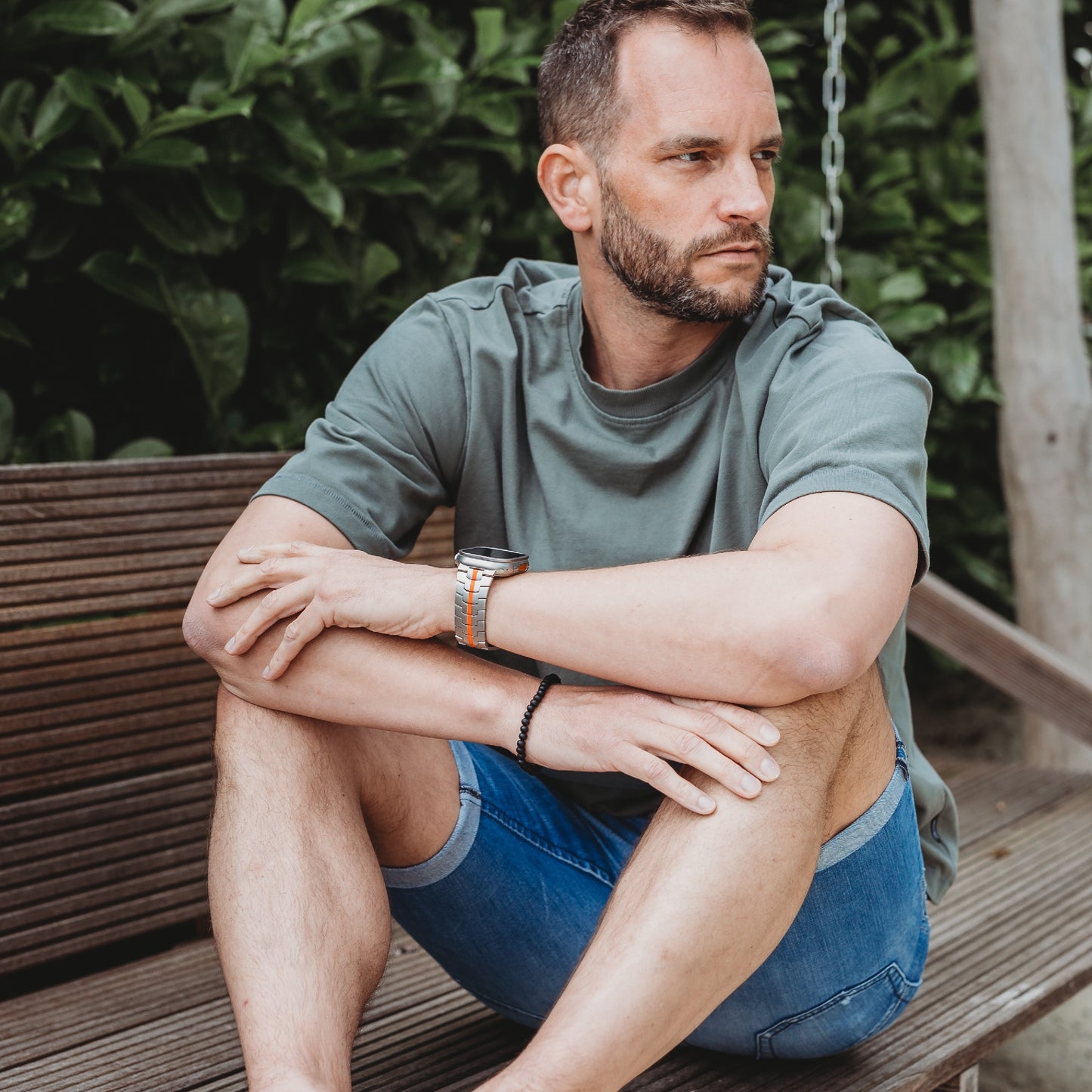Ein Mann mit kurzen Haaren und Bart sitzt im Schneidersitz auf einer Holzbank im Freien. Er trägt ein graues T-Shirt, blaue Shorts und ein Arktisband Apple Watch Ultra Armband TITANIUM F-15. Im Hintergrund sind grüne Pflanzen zu sehen.