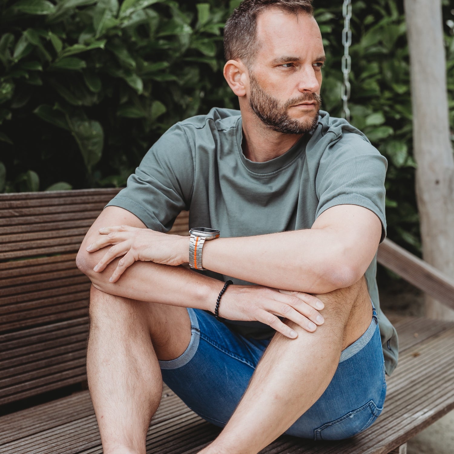 Ein Mann mit kurzen Haaren und Bart sitzt im Freien auf einer Holzbank. Er trägt ein grünes T-Shirt, Jeansshorts und das arktisband Apple Watch TITAN Armband „Aviato Edition“ von arktisband, umgeben von üppigem grünem Laub.