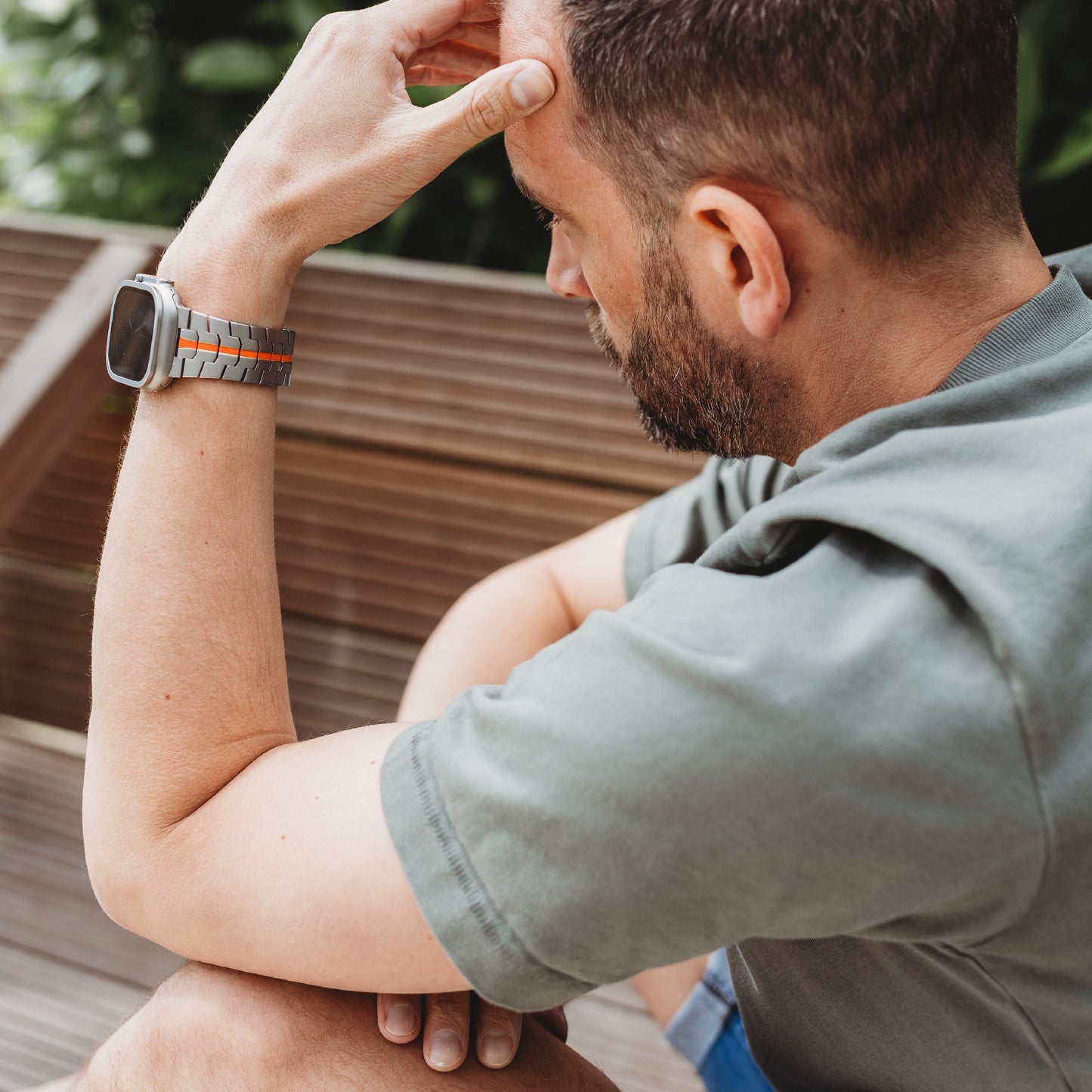 Ein Mann mit kurzen Haaren und Bart sitzt draußen auf einer Holzbank. Er trägt ein graues T-Shirt und ein Arktisband Apple Watch Ultra Armband TITANIUM F-15 von Arktisband. Er stützt seinen Ellbogen auf sein Knie und blickt nachdenklich.