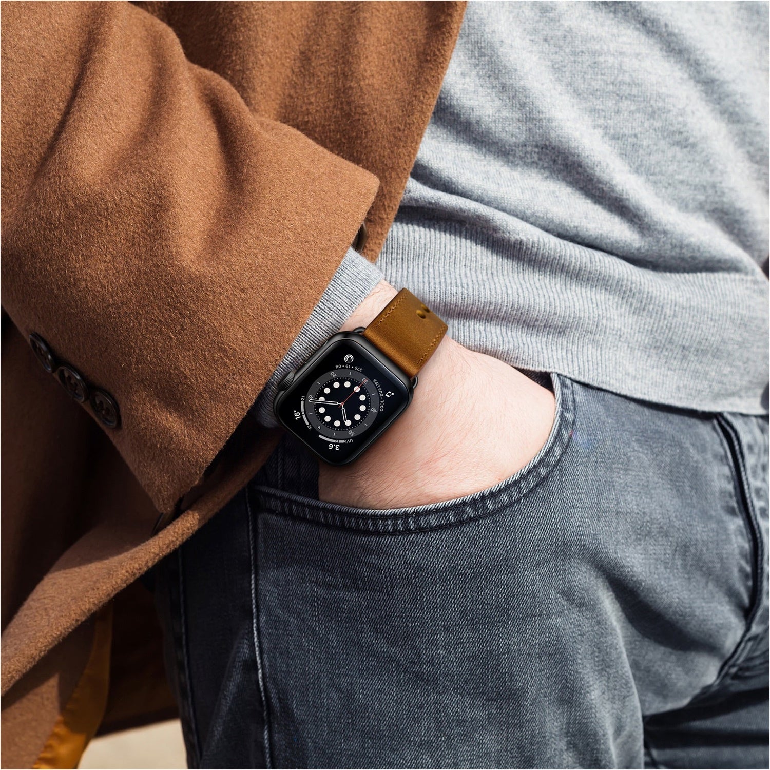 Person mit braunem Mantel, grauem Hemd, Jeans und einer Smartwatch mit braunem Lederarmband am Handgelenk.