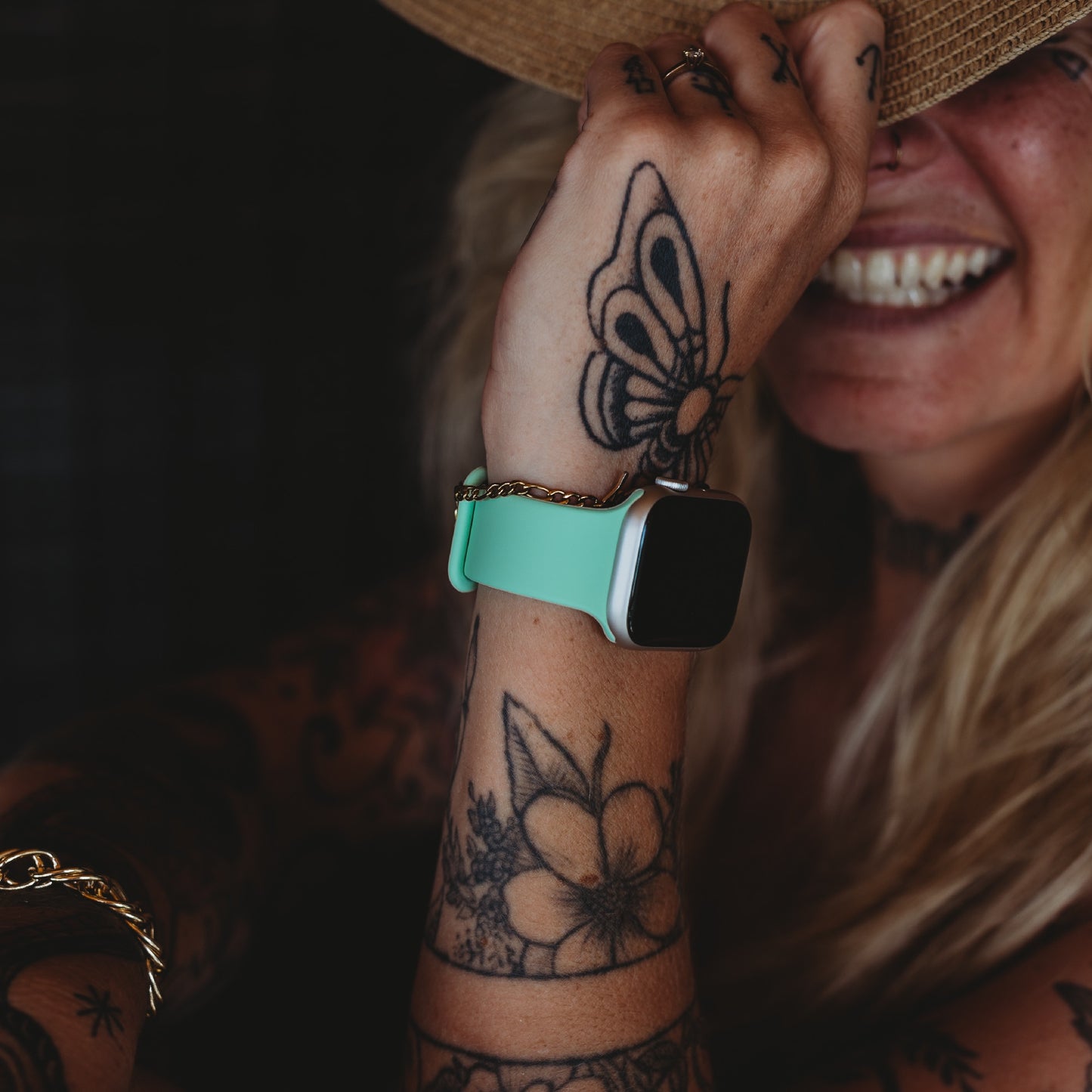Eine Person mit sichtbaren Tattoos trägt ein Arktisband Apple Watch Silikonarmband „Aqua“ und ein goldenes Kettenarmband und lächelt, während sie die Krempe eines Hutes hält, der ihr Gesicht teilweise bedeckt.